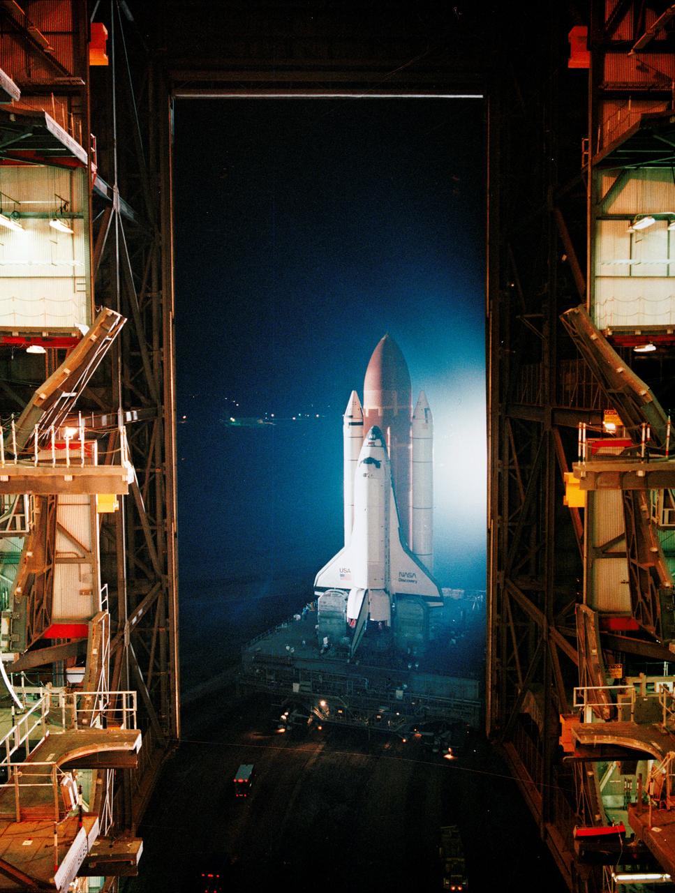 STS-26 Discovery, Orbiter Vehicle (OV) 103, mated with the external tank (ET) and solid rocket boosters (SRBs), is framed by Kennedy Space Center's (KSC's) vehicle assembly building (VAB) doorway as it rolled out. This high angle view shows OV-103 atop the mobile launch pad and crawler transporter as it begins its slow exit from VAB en route to launch complex (LC) pad 39B. The move began shortly after midnight on the nation's 212th birthday. Ceremonies marking the event were held later during daylight hours.