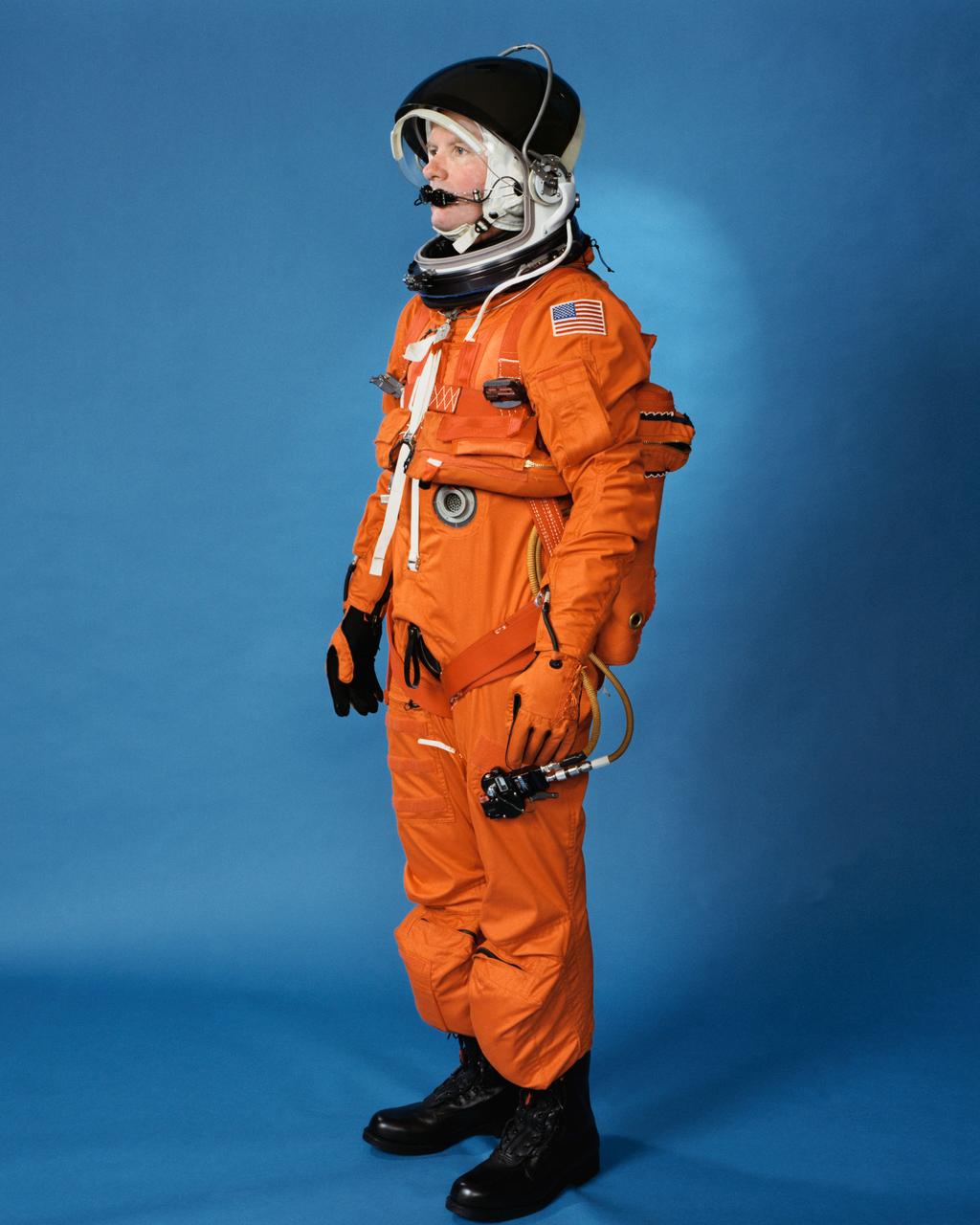 Space shuttle orange launch and entry suit (LES), a partial pressure suit, is modeled by a technician. LES was designed for STS-26, the return to flight mission, and subsequent missions. Included in the crew escape system (CES) package are launch and entry helmet (LEH) with communications carrier (COMM CAP), parachute pack and harness, life raft, life preserver unit (LPU), LES gloves, suit oxygen manifold and valves, boots, and survival gear.