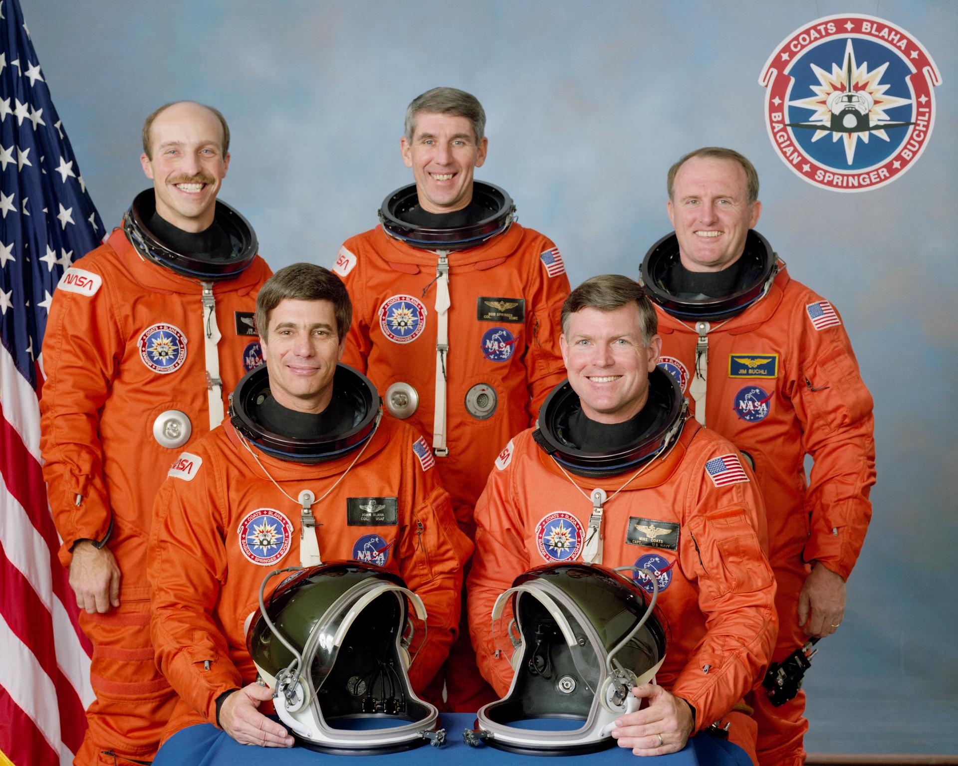 Five astronauts in orange spacesuits pose for portrait in front of US flag