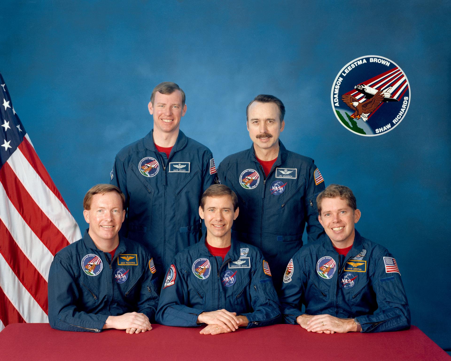 Five astronauts in blue jumpsuits posed in front of US flag.