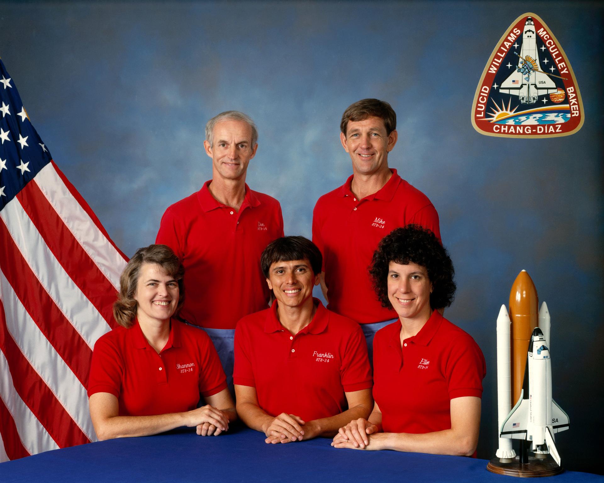 Five astronauts pose for crew portrait with model shuttle and US flag in the background.