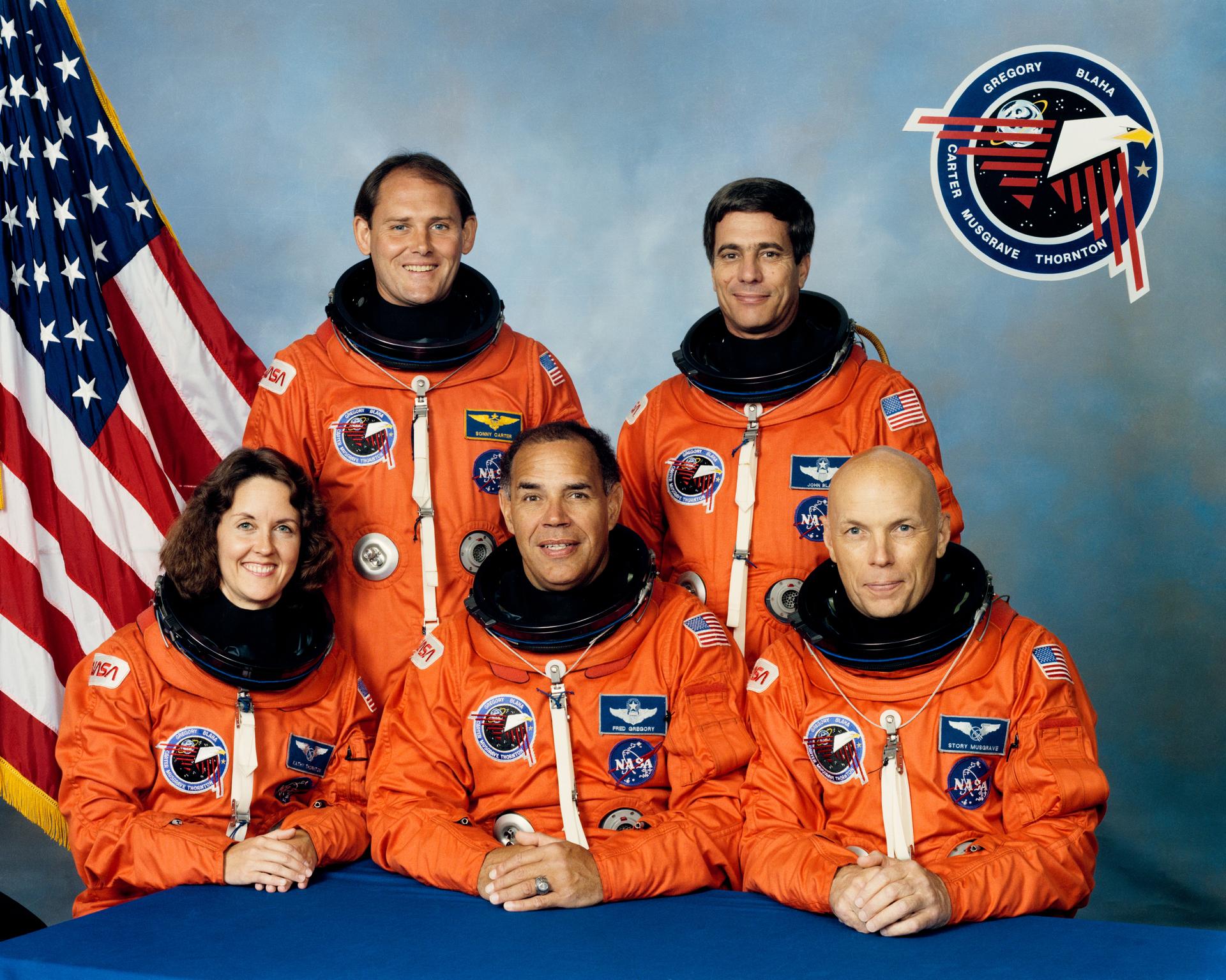 Five astronauts pose in orange space suits in front of US flag.