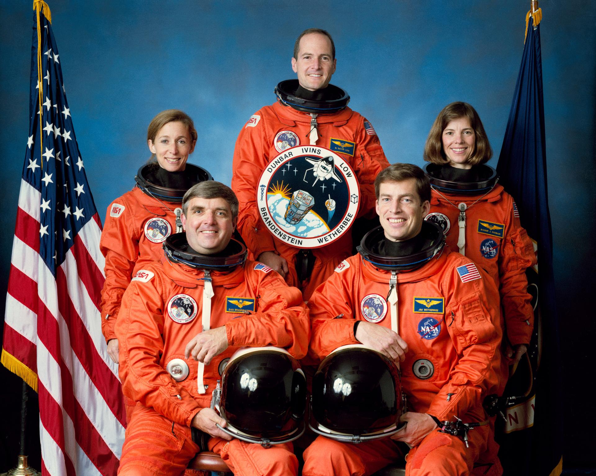 Five astronauts pose for crew photo in orange spacesuits in front of US flag.