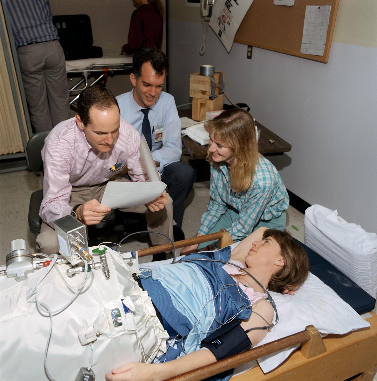STS-32 crewmembers test the inflight lower body negative pressure (LBNP) device. Mission Specialist (MS) Bonnie J. Dunbar (lying down) inside the cylindrical LBNP device prepares for testing as principal investigator Dr. John Charles, a cardiovascular scientist in JSC's Space Biomedical Research Institute, and Michele Jones, a KRUG International biomedical engineer, review procedures with MS G. David Low. The inflight LBNP will be part of detailed supplementary objective (DSO) 0478. Photo taken by JSC photographer Jack Jacob.