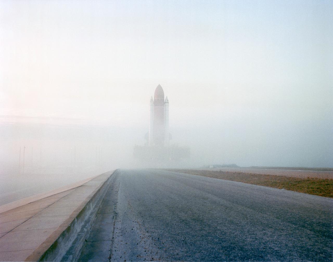 STS-32 Columbia, Orbiter Vehicle (OV) 102, rolls through the morning's foggy mist atop the mobile launcher platform and crawler transporter to Kennedy Space Center (KSC) Launch Complex (LC) Pad 39A. OV-102's wings appear on either side of the two solid rocket boosters (SRBs) and external tank (ET). Rollout from the Vehicle Assembly Building (VAB) began at 2:32 am Eastern Standard Time (EST), and OV-102 was on the pad pedestals about 8 hours later. This marks the first time a Space Shuttle has been at LC Pad 39A since 01-12-85 when OV-102 was launched on mission 61C. View provided by KSC with alternate number KSC-89PC-1259.