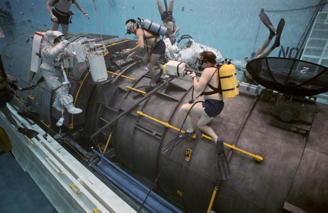 STS-31 Mission Specialist (MS) Bruce McCandless II (left), wearing an extravehicular mobility unit (EMU), maneuvers his way around a mockup of the remote manipulator system (RMS) end effector during an underwater simulation in JSC's Weightless Environment Training Facility (WETF) Bldg 29 pool. The end effector is attached to a grapple fixture on the Hubble Space Telescope (HST) mockup. As McCandless performs contingency extravehicular activity (EVA) procedures, fellow crewmember MS Kathryn D. Sullivan, in EMU, works on the opposite side of the HST mockup, and SCUBA-equipped divers monitor the activity. Though no EVA is planned for STS-31, the two crewmembers train for contingencies that would necessitate leaving the shirt sleeve environment of Discovery's, Orbiter Vehicle (OV) 103's, crew cabin and performing chores with the HST payload or related hardware in the payload bay (PLB).