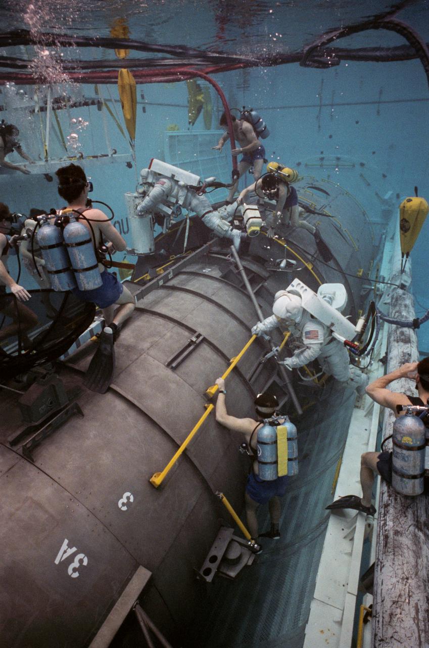 This overall view shows STS-31 Mission Specialist (MS) Bruce McCandless II (left) and MS Kathryn D. Sullivan making a practice space walk in JSC's Weightless Environment Training Facility (WETF) Bldg 29 pool. McCandless works with a mockup of the remote manipulator system (RMS) end effector which is attached to a grapple fixture on the Hubble Space Telescope (HST) mockup. Sullivan manipulates HST hardware on the Support System Module (SSM) forward shell. SCUBA-equipped divers monitor the extravehicular mobility unit (EMU) suited crewmembers during this simulated extravehicular activity (EVA). No EVA is planned for the Hubble Space Telescope (HST) deployment, but the duo has trained for contingencies which might arise during the STS-31 mission aboard Discovery, Orbiter Vehicle (OV) 103. Photo taken by NASA JSC photographer Sheri Dunnette.