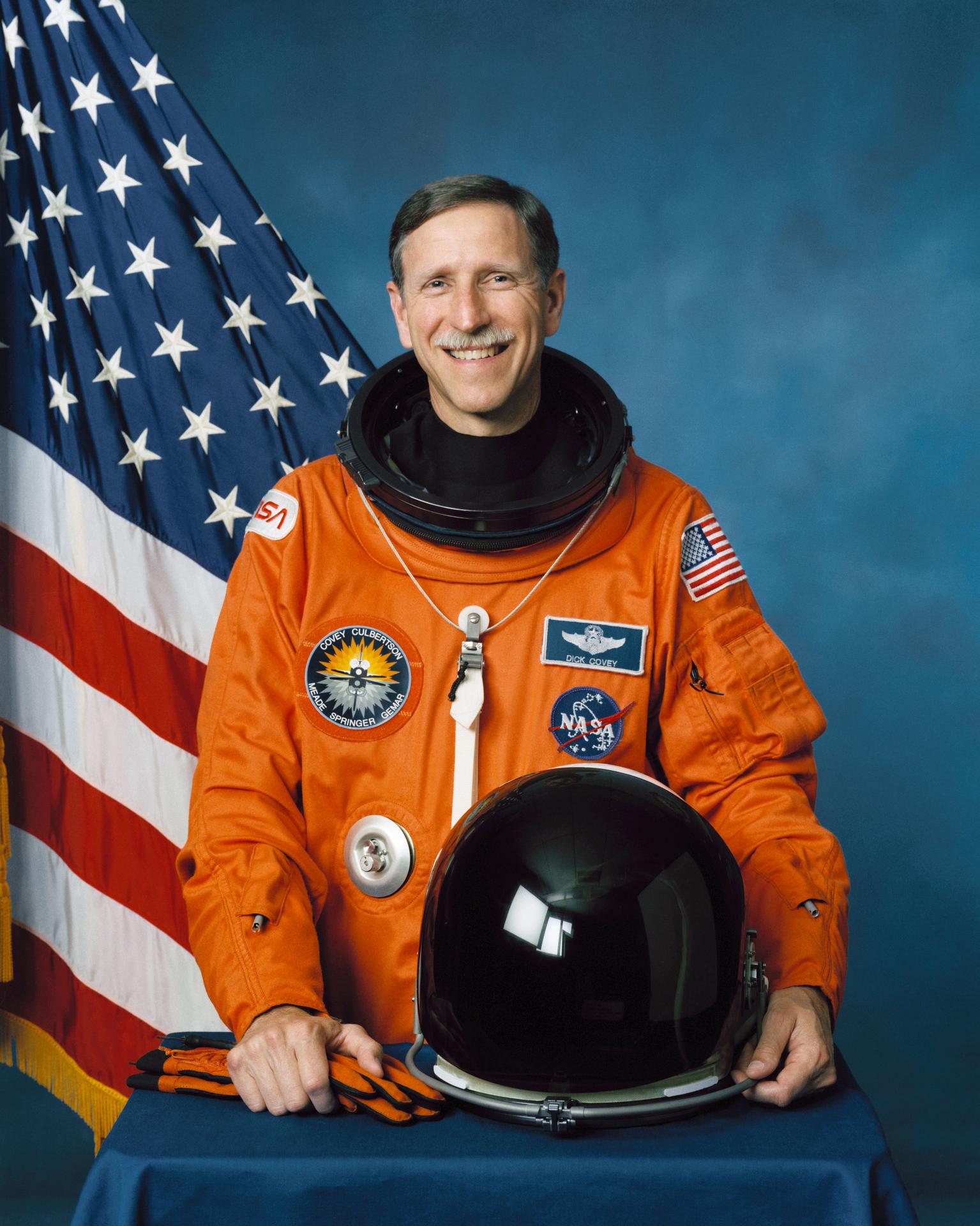 A man smiles, wearing a spacesuit, and stands in front of the American flag. He has his helmet off and sitting on table before him.