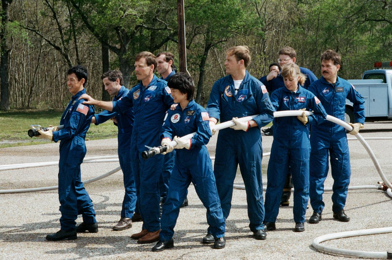 STS-47 Endeavour, Orbiter Vehicle (OV) 105, crewmembers led by Commander Robert L. Gibson (center) prepare to extinguish a blaze in JSC's Fire Training Pit. Lined up along the water hoses are: (on left) Payload Specialist Mamoru Mohri, holding the hose nozzle, followed by Mission Specialist (MS) Jerome Apt, and Pilot Curtis L. Brown, Jr; and (on right) backup Payload Specialist Chiaki Naito-Mukai, holding the hose nozzle, followed by MS and Payload Commander (PLC) Mark C. Lee, MS N. Jan Davis, and backup Payload Specialist Stan Koszelak. A veteran fire fighter and the instructor, positioned between the two hoses, looks on. Mohri and Mukai represent Japan's National Space Development Agency (NASDA). The Fire Training Pit is located across from the Gilruth Center Bldg 207.