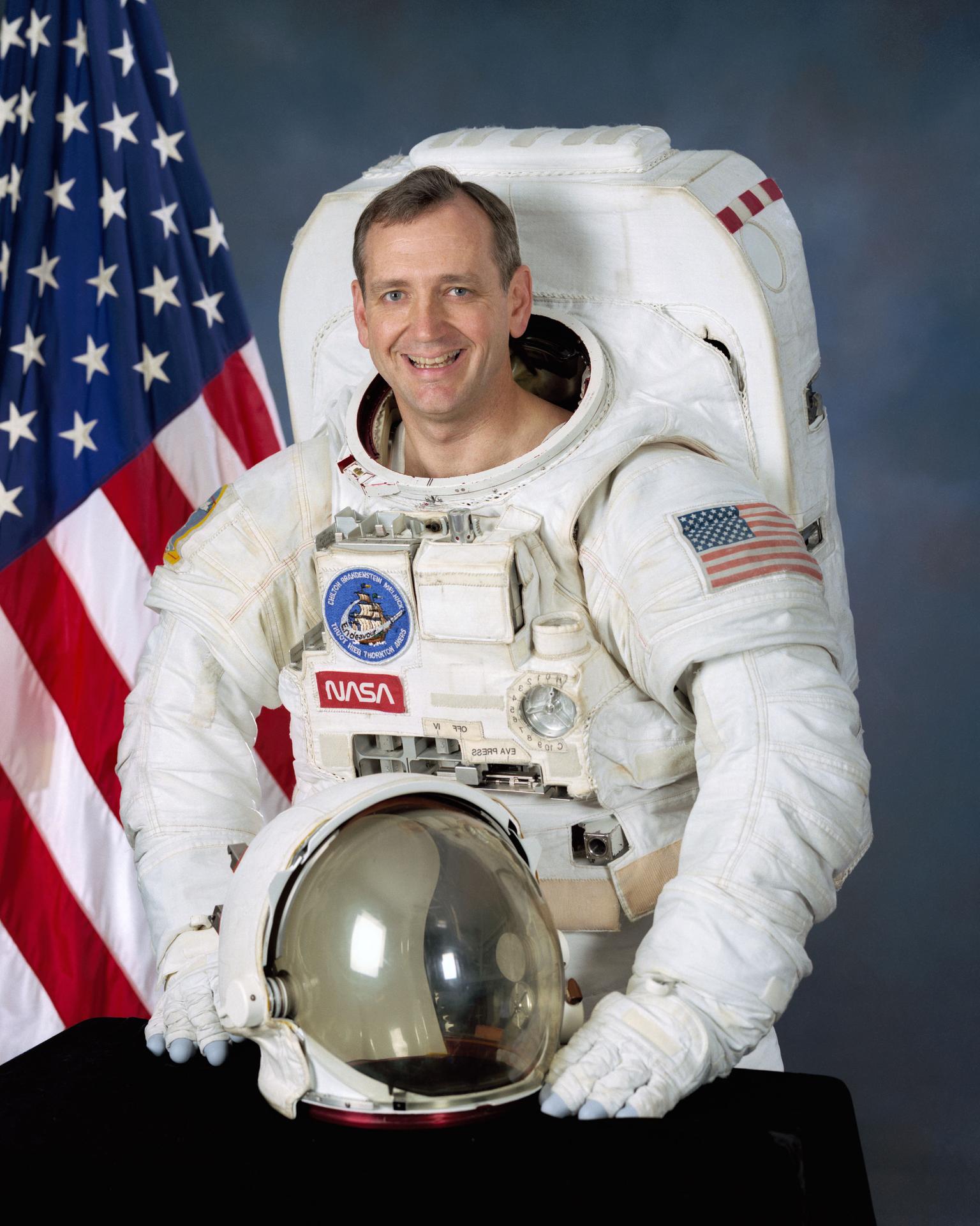 A man sits before the American flag in an astronaut suite, with his helmet off