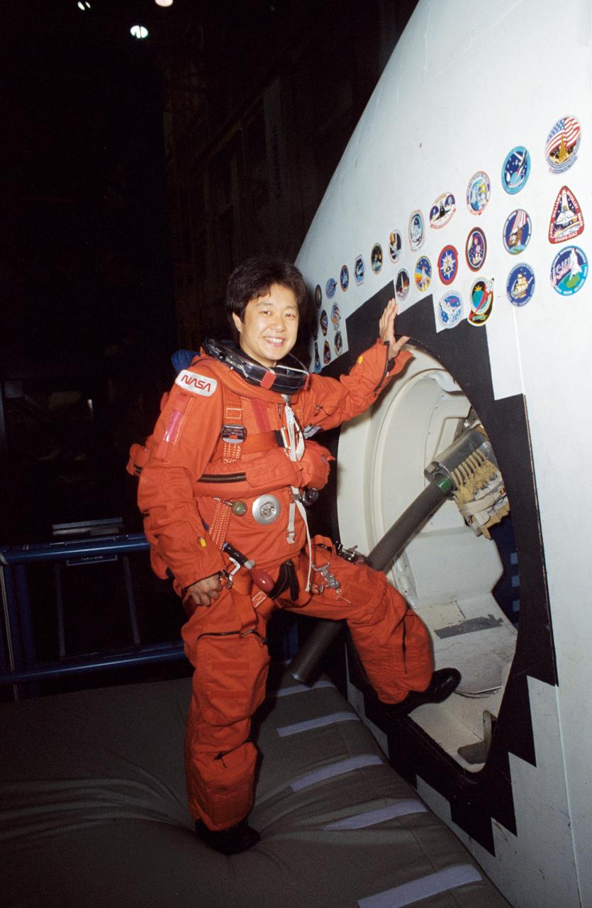 STS-65 Japanese Payload Specialist Chiaki Mukai takes a break from training at the Johnson Space Center (JSC). Wearing a training version of the orange launch and entry suit (LES), Mukai stands at the crew compartment trainer (CCT) side hatch in the Mockup and Integration Laboratory (MAIL) Bldg 9NE. Note the crew escape system (CES) pole device extending out the side hatch which would accommodate crewmembers in bailout from a troubled spacecraft. Mukai represents the National Space Development Agency (NASDA) of Japan and will serve as a payload specialist aboard Columbia, Orbiter Vehicle (OV) 102, during the STS-65 International Microgravity Laboratory 2 (IML-2) mission.