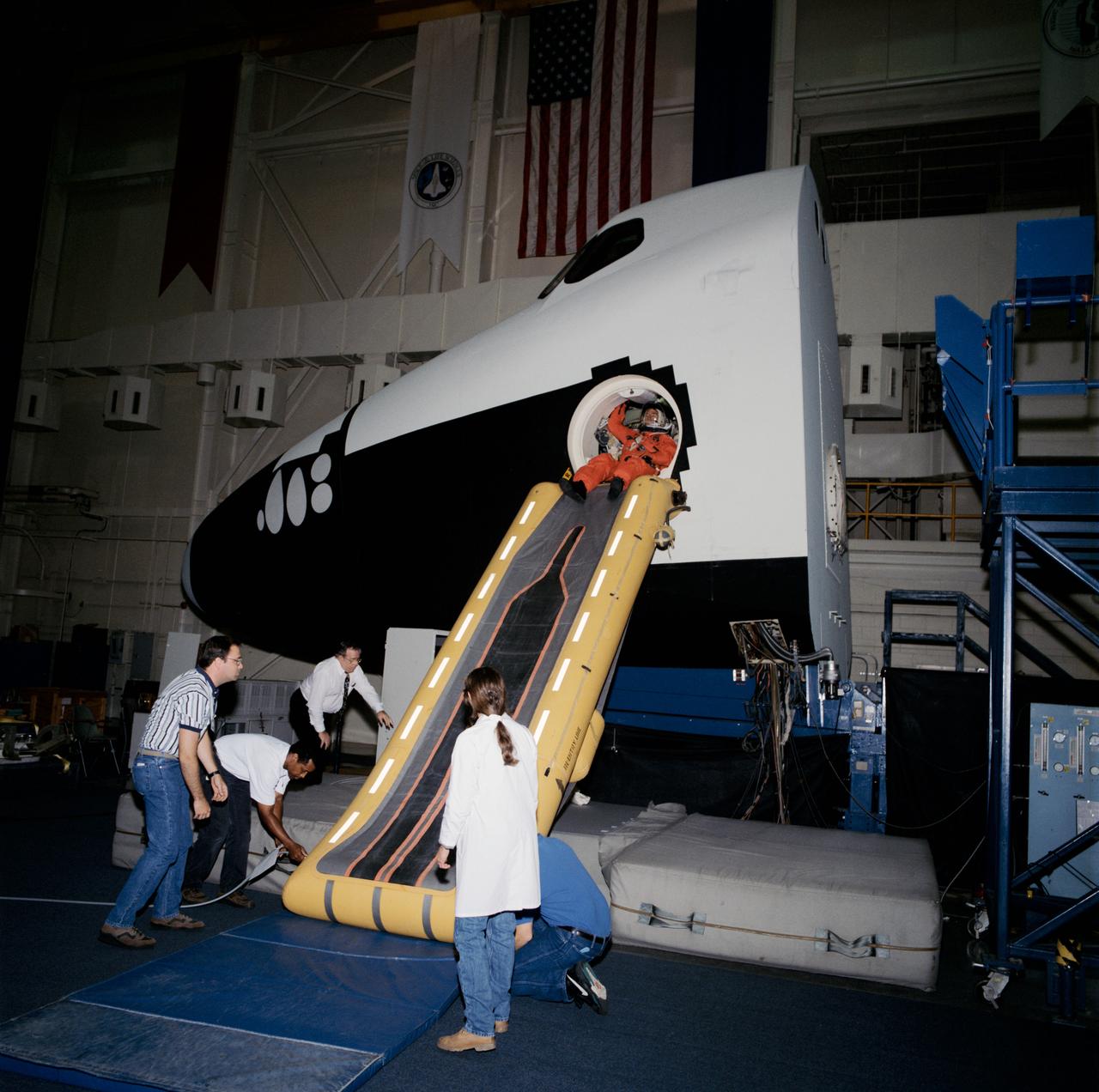 View of the STS-73 crew using the escape slide in the Crew Compartment Trainer (CCT) at bldg 9A. The crew is seen in their launch and entry suits at the top of the slide with trainers at the bottom.