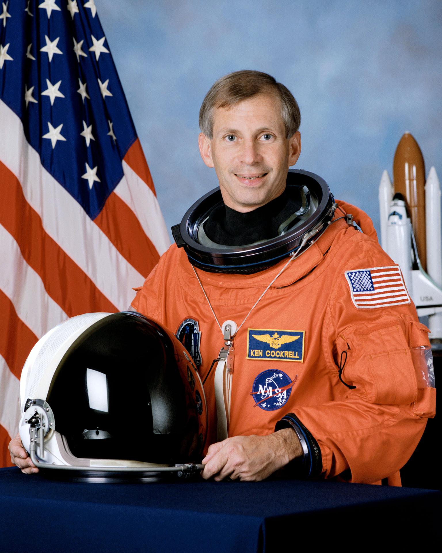 A man wearing a spacesuit smiles and stands in front of the American flag. He is holding his helmet in front of himself.