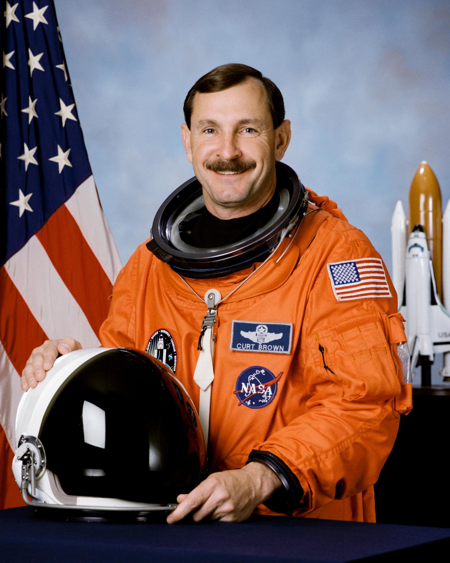 A man wearing a spacesuit smiles and stands in front of the American flag. He is holding his helmet in front of himself.