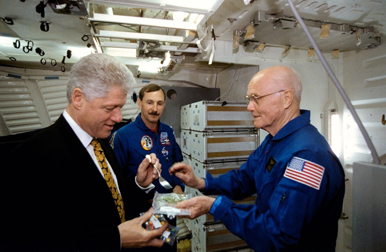 S98-05017 (14 April 1998) --- President Bill Clinton prepares to use a fork to sample some space food while visiting NASA's Johnson Space Center (JSC). Holding the food packet is U.S. Sen. John H. Glenn Jr. (D.-Ohio), currently in training at JSC as a payload specialist for a flight scheduled later this year aboard the Space Shuttle Discovery. Looking on is astronaut Curtis L. Brown Jr., STS-95 commander. The picture was taken in the full fuselage trainer (FFT). Photo Credit: Joe McNally, National Geographic, for NASA