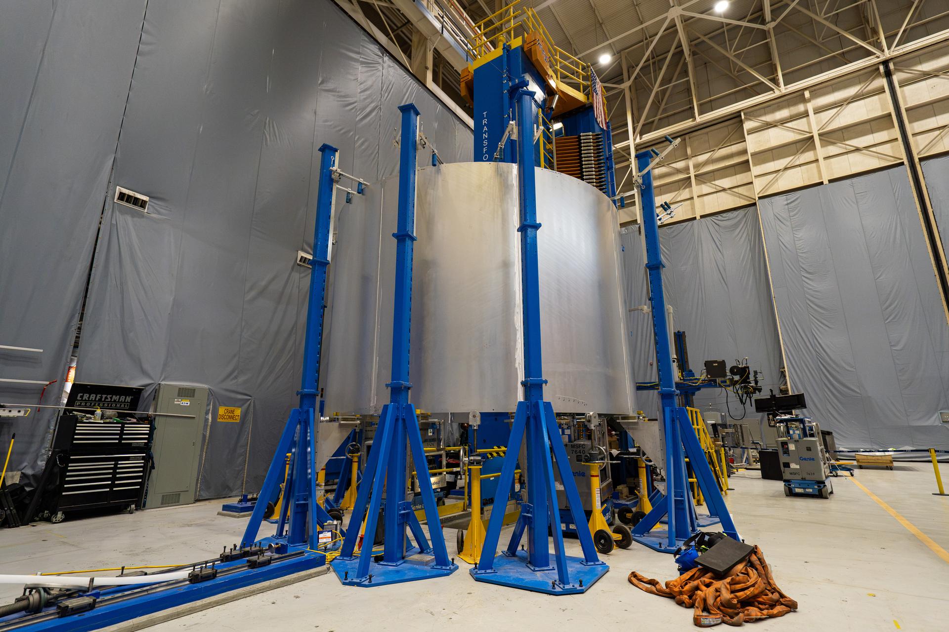 These images show the spacer barrel for NASA's SLS (Space Launch System), which will be used for the Artemis III mission. Inside a high-bay manufacturing facility at NASA's Marshall Space Flight Center in Huntsville, Alabama, the large silver barrel stands vertically after technicians completed the eighth and final friction stir weld joining its eight panels. Blue and yellow platforms, scaffolding, and specialized welding equipment surround the large piece of hardware.