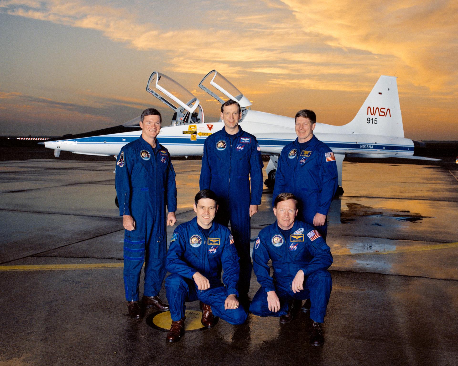 Five men in blue jumpsuits pose in front of shuttle.