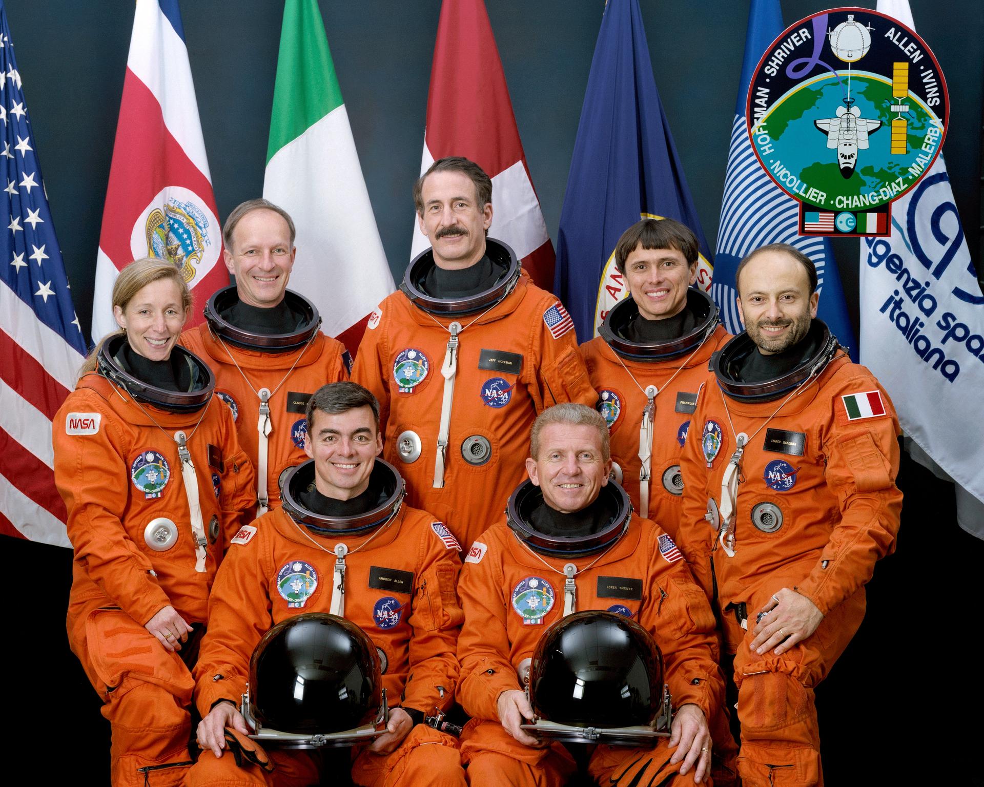 Five men and one woman in spacesuits pose for crew photo in front of flags.