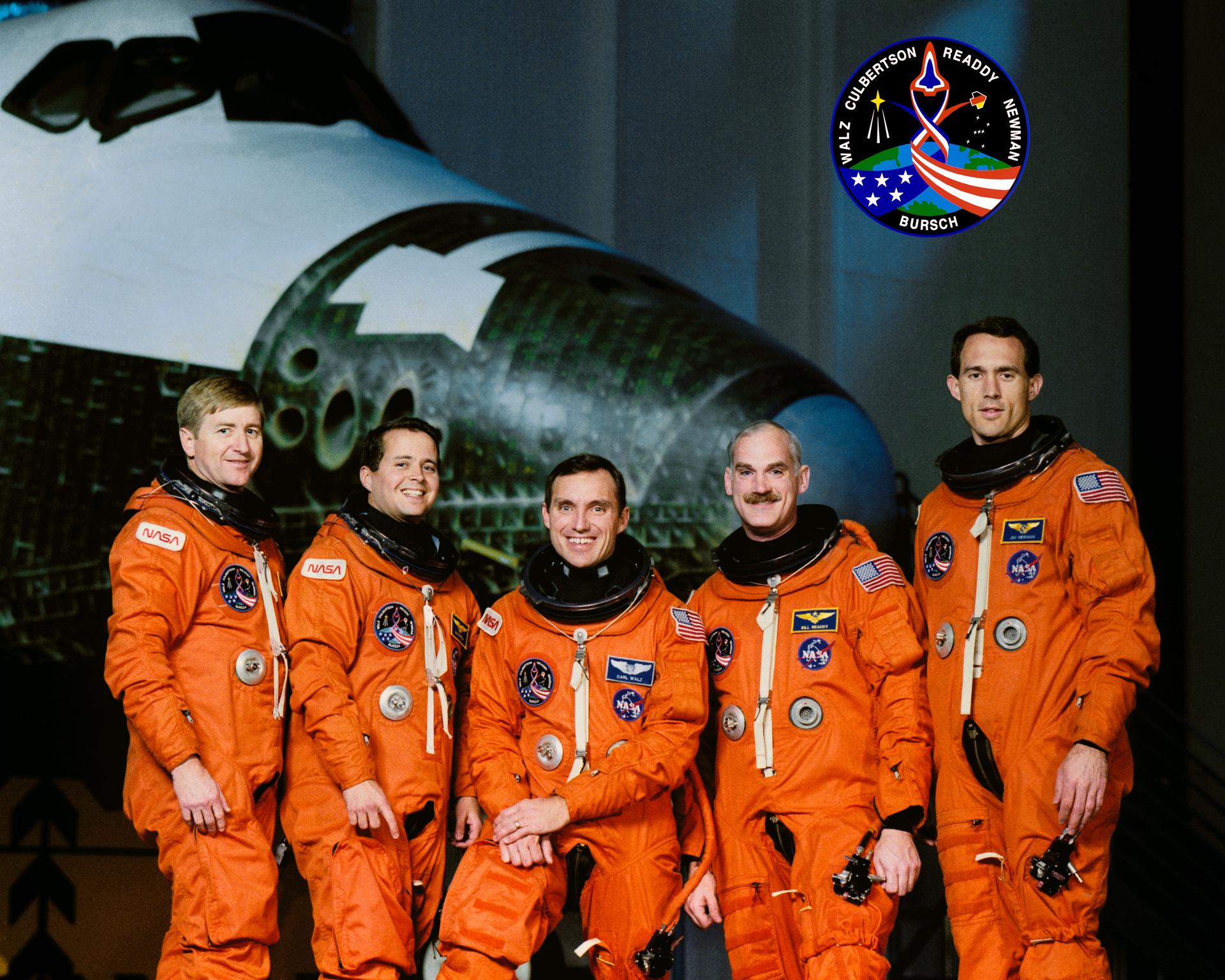 The five crew members of the STS-51 mission pose for a crew portrait in their orange spacesuits in front of space shuttle.