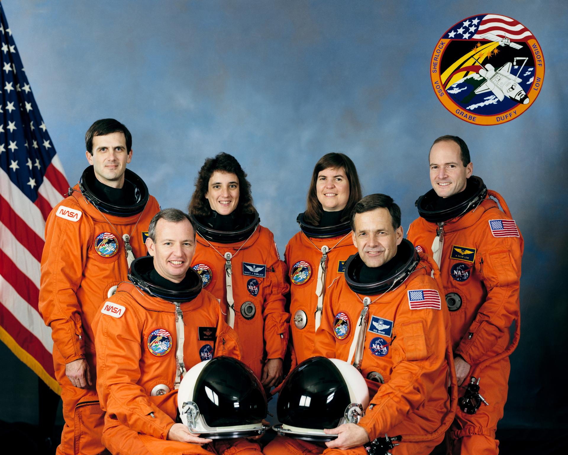 Four men and two women in orange spacesuits pose for crew photo in front of American flag.