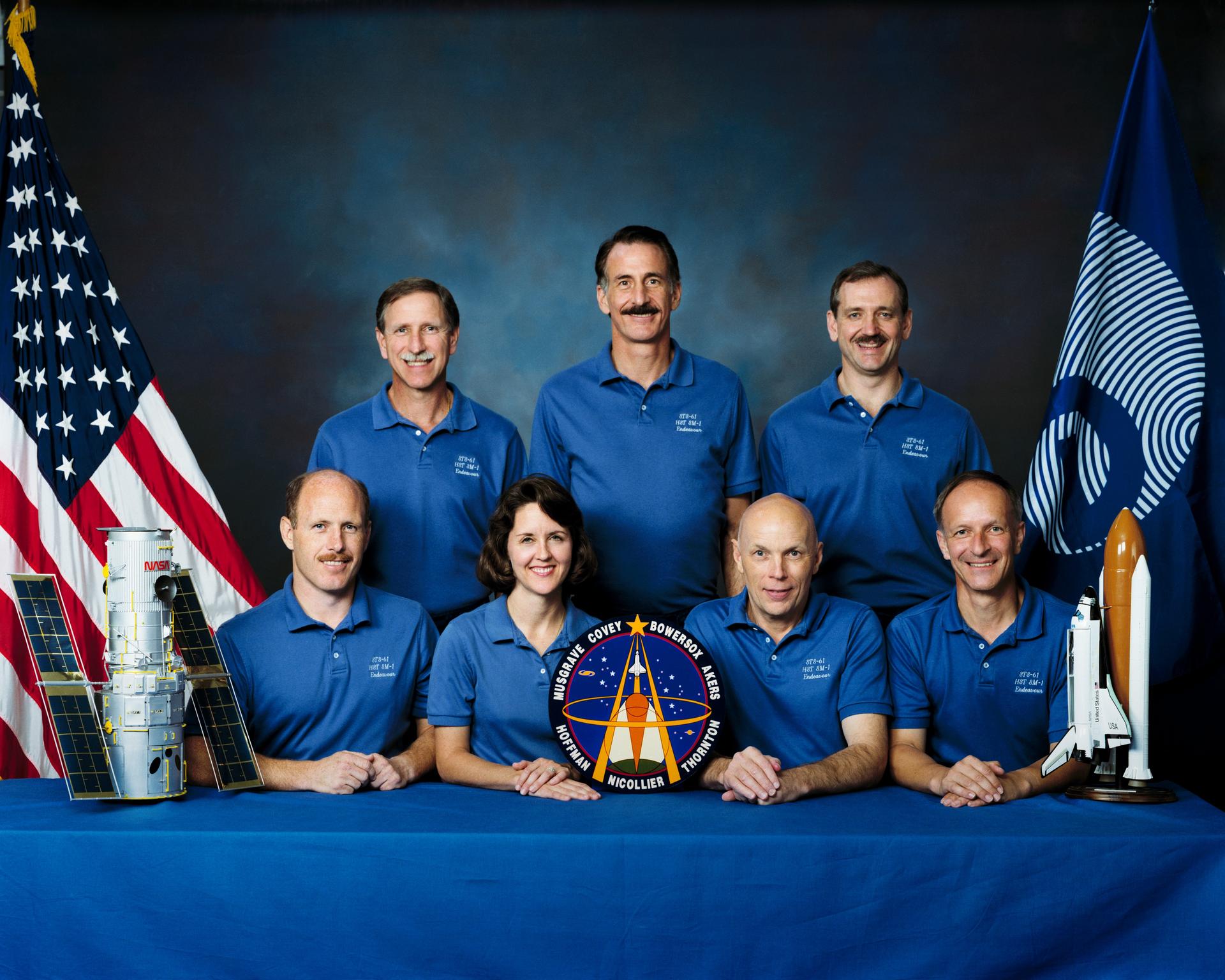 STS-61 crew portrait with a model of the Hubble on the left in front, and a model of the Shuttle on the right. The American flag and the ESA flag are in the background.