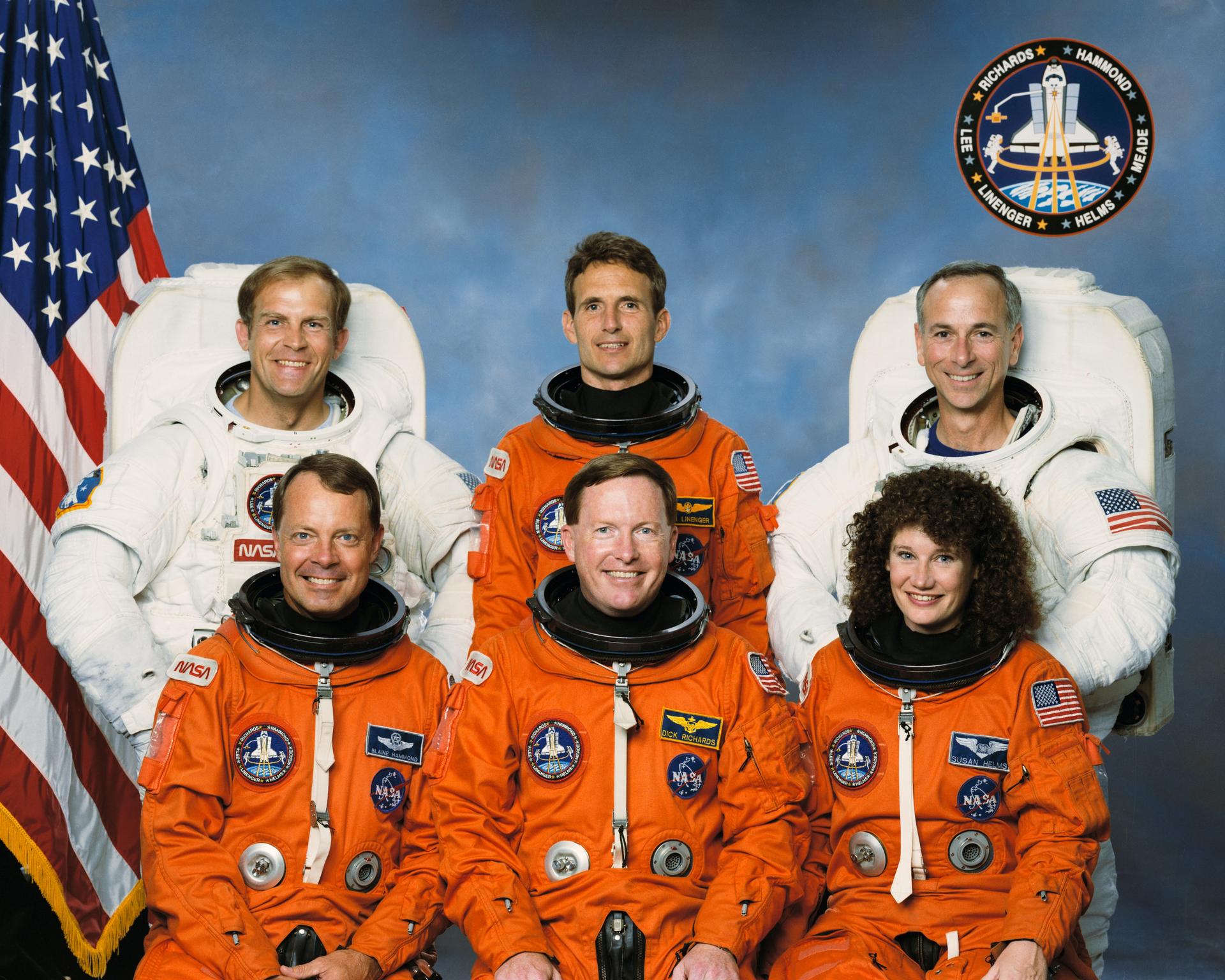 Four astronauts in orange suits and two in white spacesuits pose for crew photo in front of American flag.