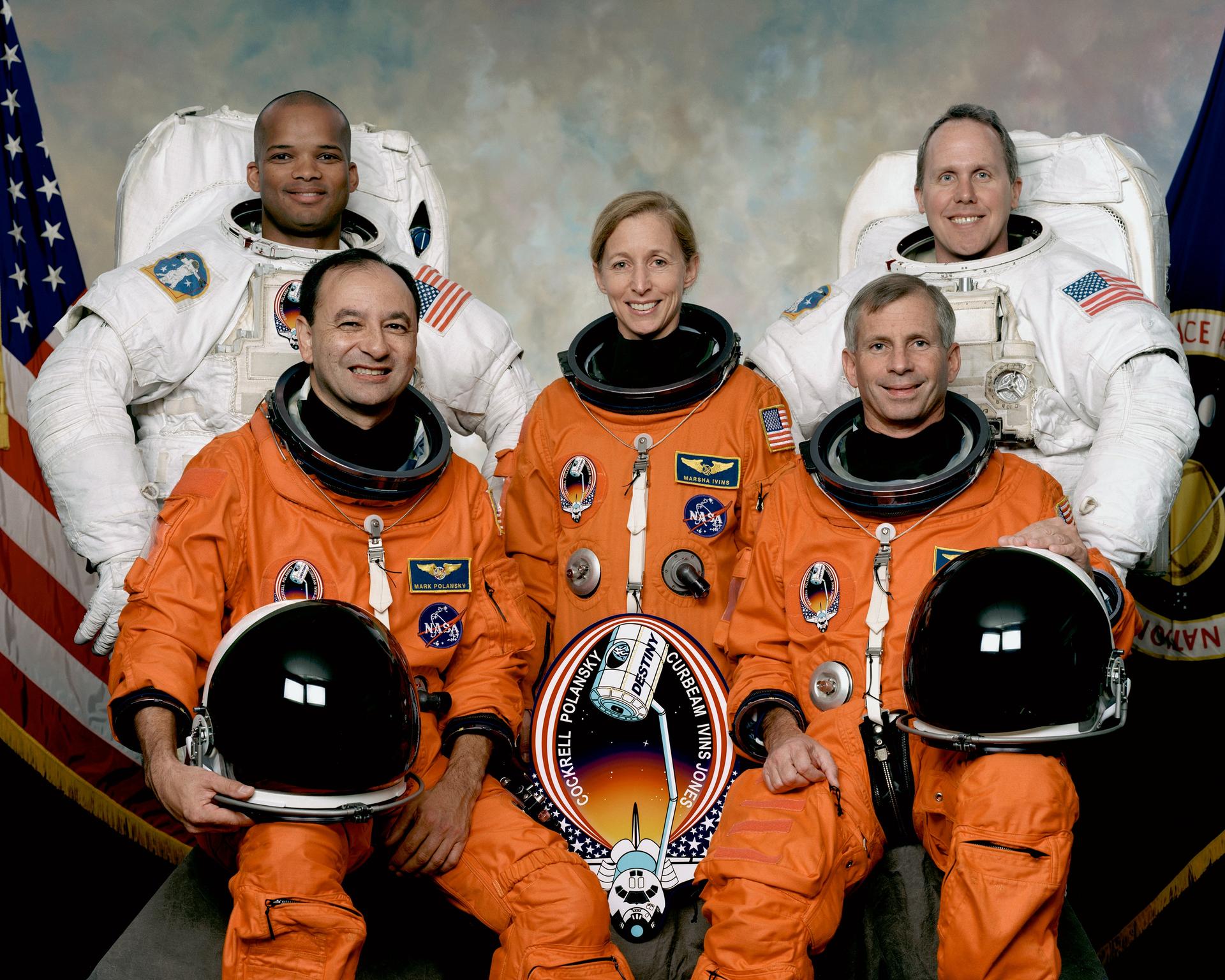 Five astronauts in orange and white spacesuits pose for portrait in front of flags