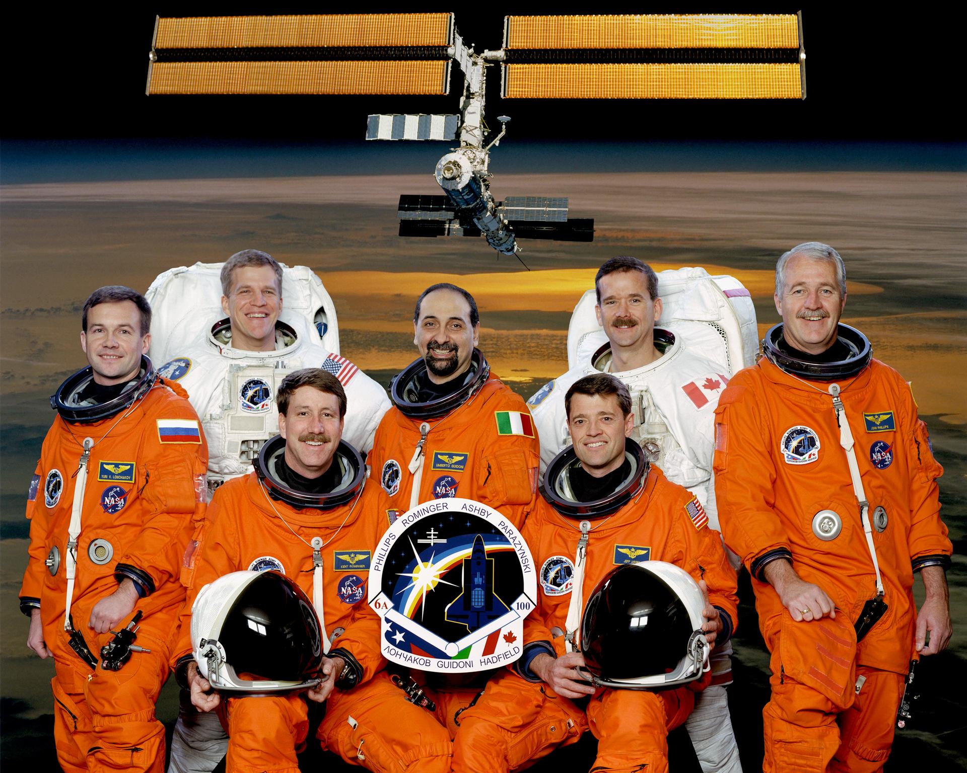 Seven astronauts pose for portrait in orange and white spacesuits with the International Space Station in the background