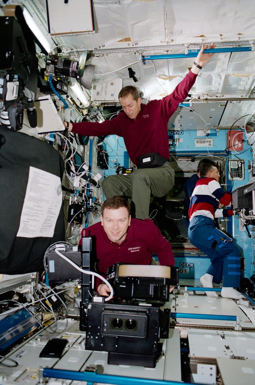 STS102-317-027 (8-21 March 2001)--- Onboard the Destiny laboratory on the International Space Station (ISS), astronaut James D. Wetherbee, mission commander, is photographed in a surfing position on astronaut James M. Kelly, pilot, while he is loading the IMAX camera. Astronaut James S. Voss, Expedition Two flight engineer is visible working in the background.