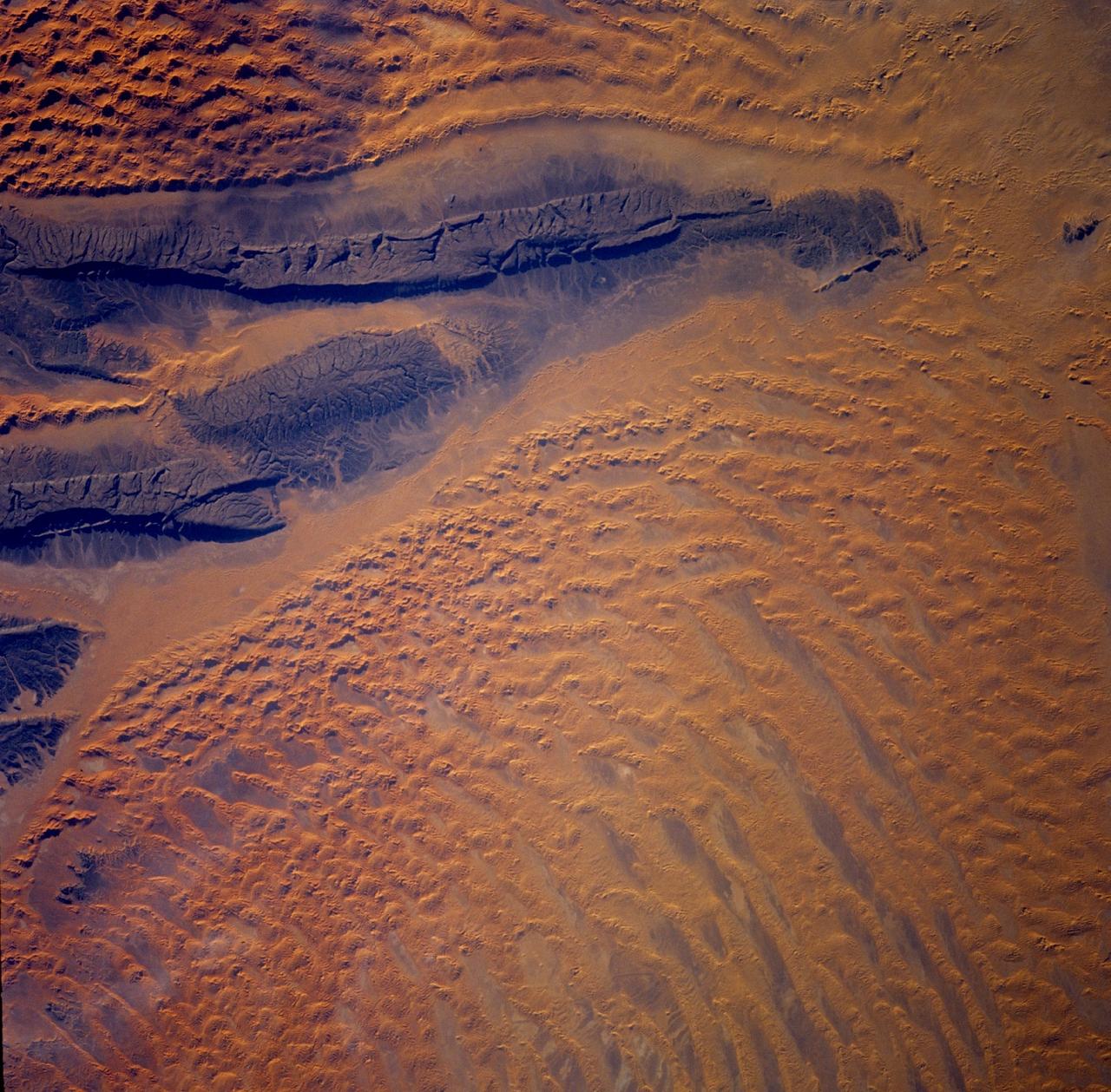 STS103-728-022 (19-27 December 1999)--- One of the astronauts aboard the Earth-orbiting Space Shuttle Discovery used a handheld 70mm camera to photograph the Tifernine dunes (note, the  dunes are below the &quot;beak&quot; of sandstone rock).  According to NASA scientists studying the STS-103 photo collection, the dunes were created when the dark sandstone rocks trapped sand.  Winds, they continued, then piled the sand into dunes up to 457.2 m (1,500 ft).  The color of the sandstone is due to a desert varnish, the scientists reported.  The varnish is composed of manganese, iron oxides, hydroxides, and clay minerals, they said.