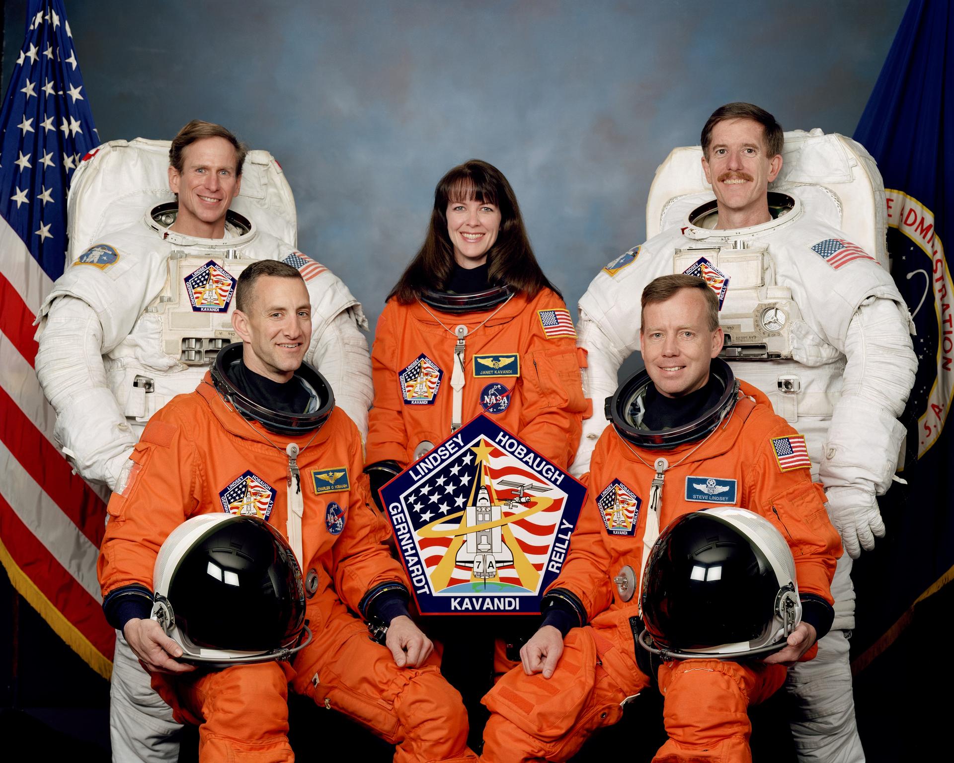 Five astronauts pose for portrait in orange and white spacesuits with flags in background