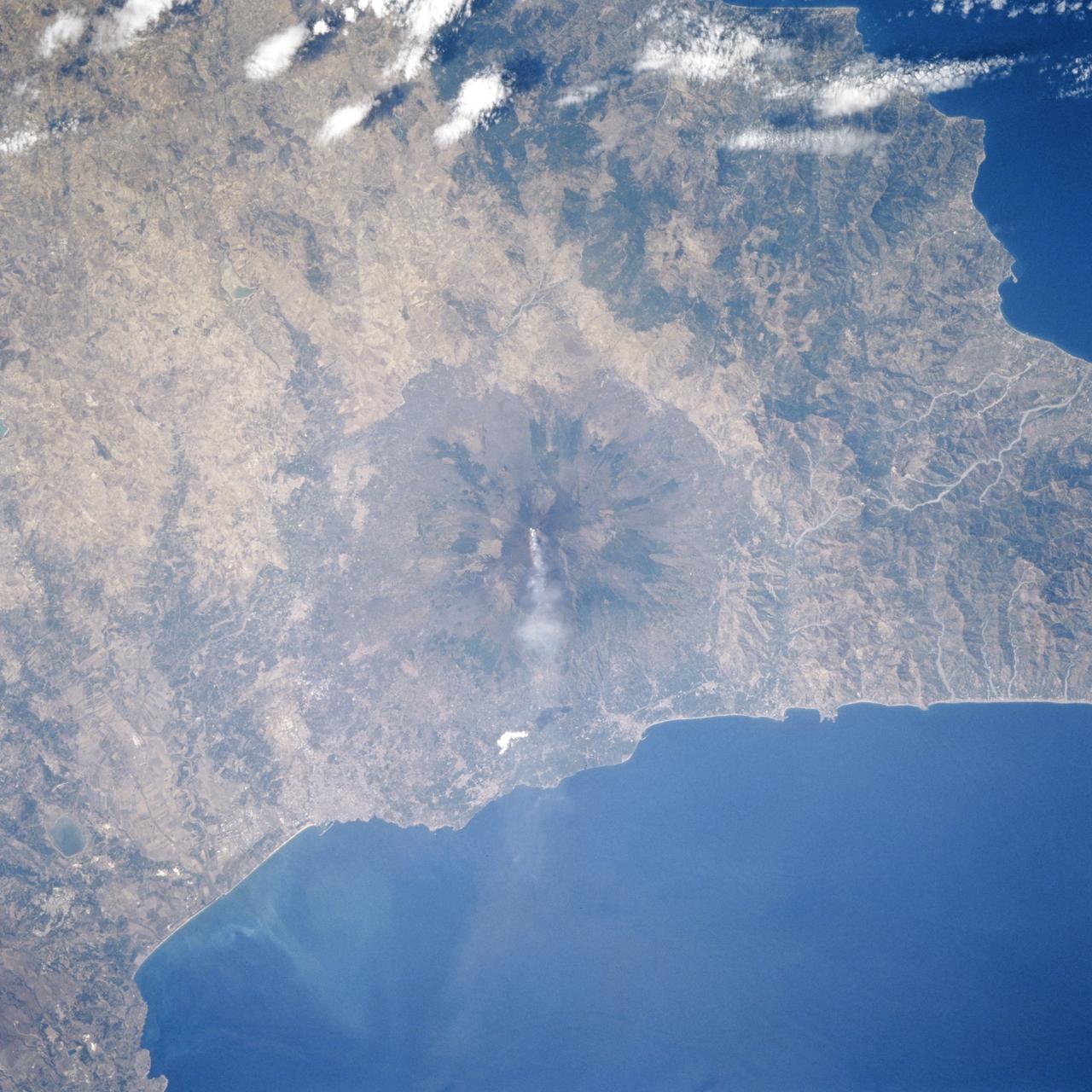 STS106-706-055 (8-20 September 2000) --- One of the STS-106 crew members on board the Space Shuttle Atlantis used a handheld 70mm camera to photograph this image of the Etna Volcano on Sicily. Mt. Etna is known as the volcano with the longest record of volcanic activity with the first being in 1500 BC. It has erupted many times since then and is almost continuously venting gas and steam, as shown in this view. Astronauts can almost be assured of seeing some venting whenever they fly in orbit. Mt. Etna appears as a cone with an almost circular base in this near-vertical view. The Salso River winds around the western and southern flanks of the volcano. The city of Catania appears as a diffuse gray patch at the foot of the volcano where the river meets the Mediterranean Sea. Mt. Etna has a complex of cones at its summit, which is nearly 3300 meters above sea level. Its slopes are a patchwork of colors, the darker colors being lava flows of different ages. Greens are patches of forest on slopes which have not been disrupted by lava and ash in the last few decades. Mt. Etna is a constructional landform which has been built upwards for millennia; it contrasts subtly but distinctly in this view with the surrounding lower hills which are water-eroded landforms everywhere sculpted into V-shaped valleys by the erosive power of flowing water of streams.