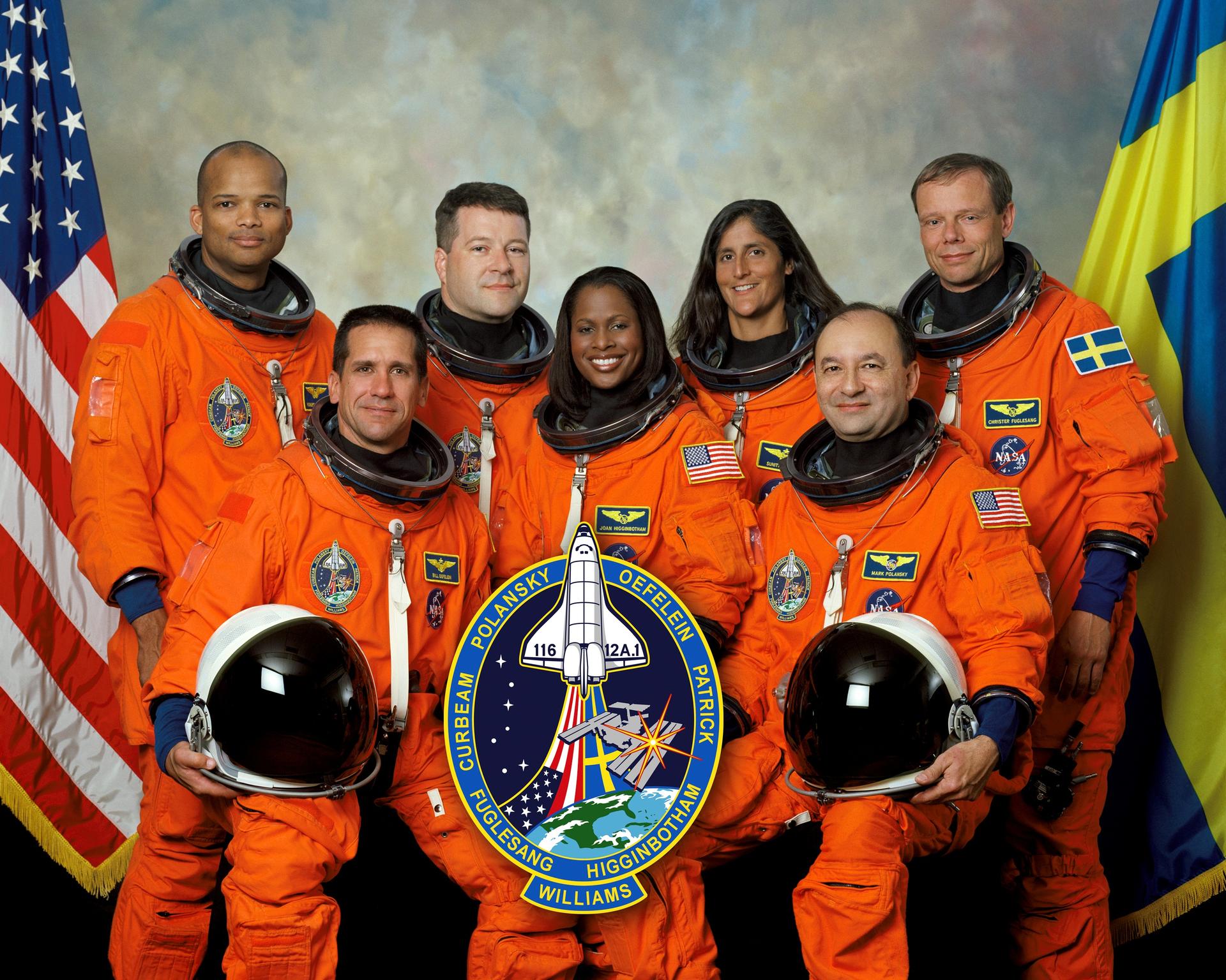 Crew members pose in orange suits in front of flags.