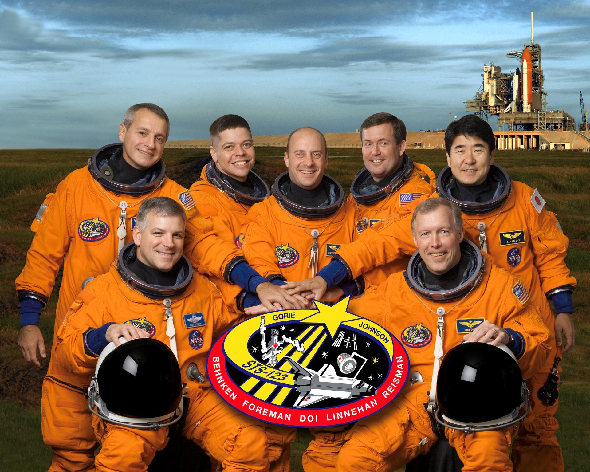 The STS-123 crew portrait shows the crew grouped together with the mission patch in front and crew members, dressed in the orange launch and entry suits, with joined hands over the patch.