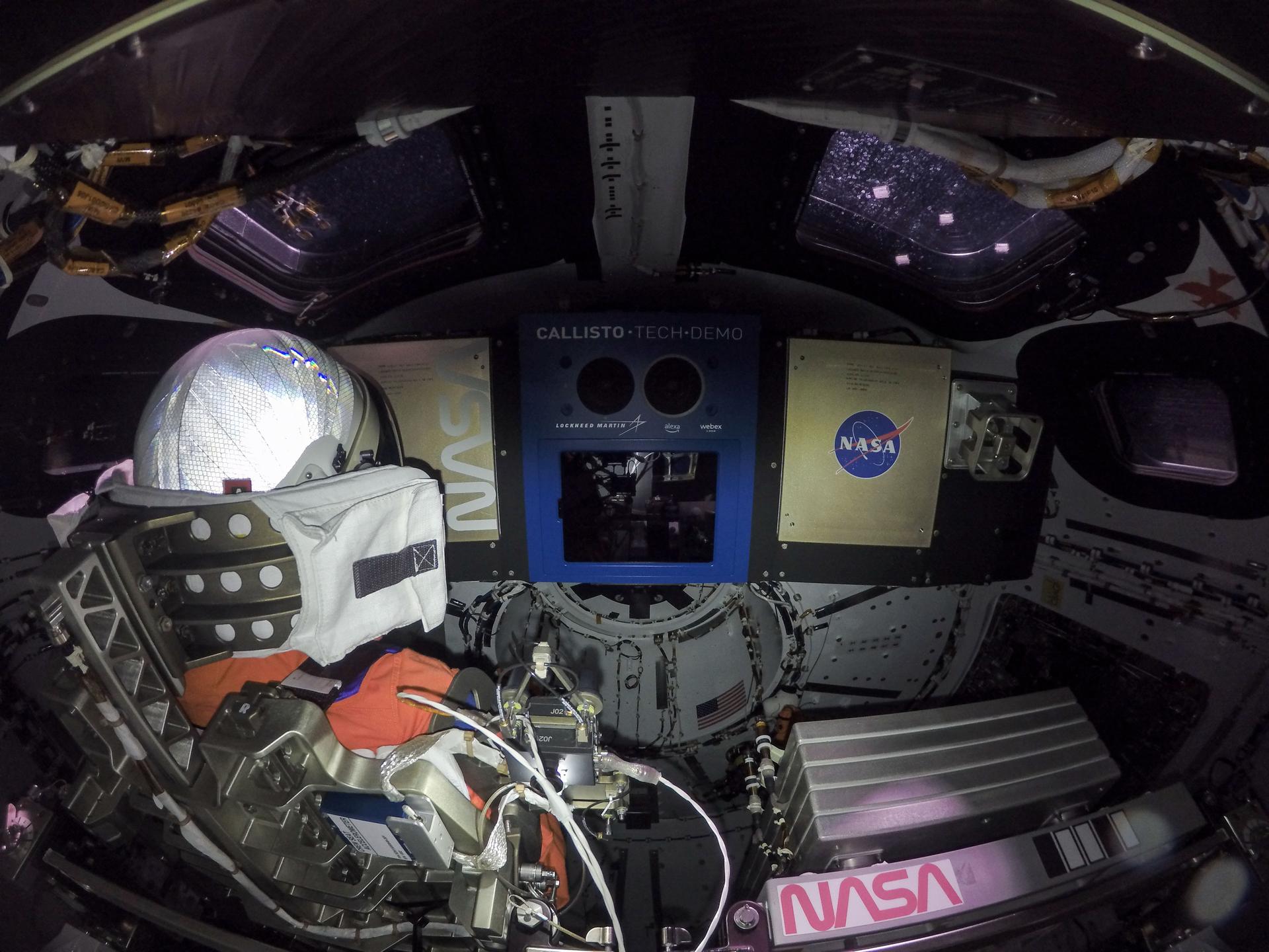 Two windows at the top of the frame showcase the darkness of space outside the crew module, while the back of Commander Moonikin Campos' white helmet is visible on the left. There is a large blue box in the center with an unlit screen -- this is the Callisto payload. A NASA "meatball" logo is to the right. Tucked away below Callisto is astronaut Snoopy, his smile and orange space suit just visible.