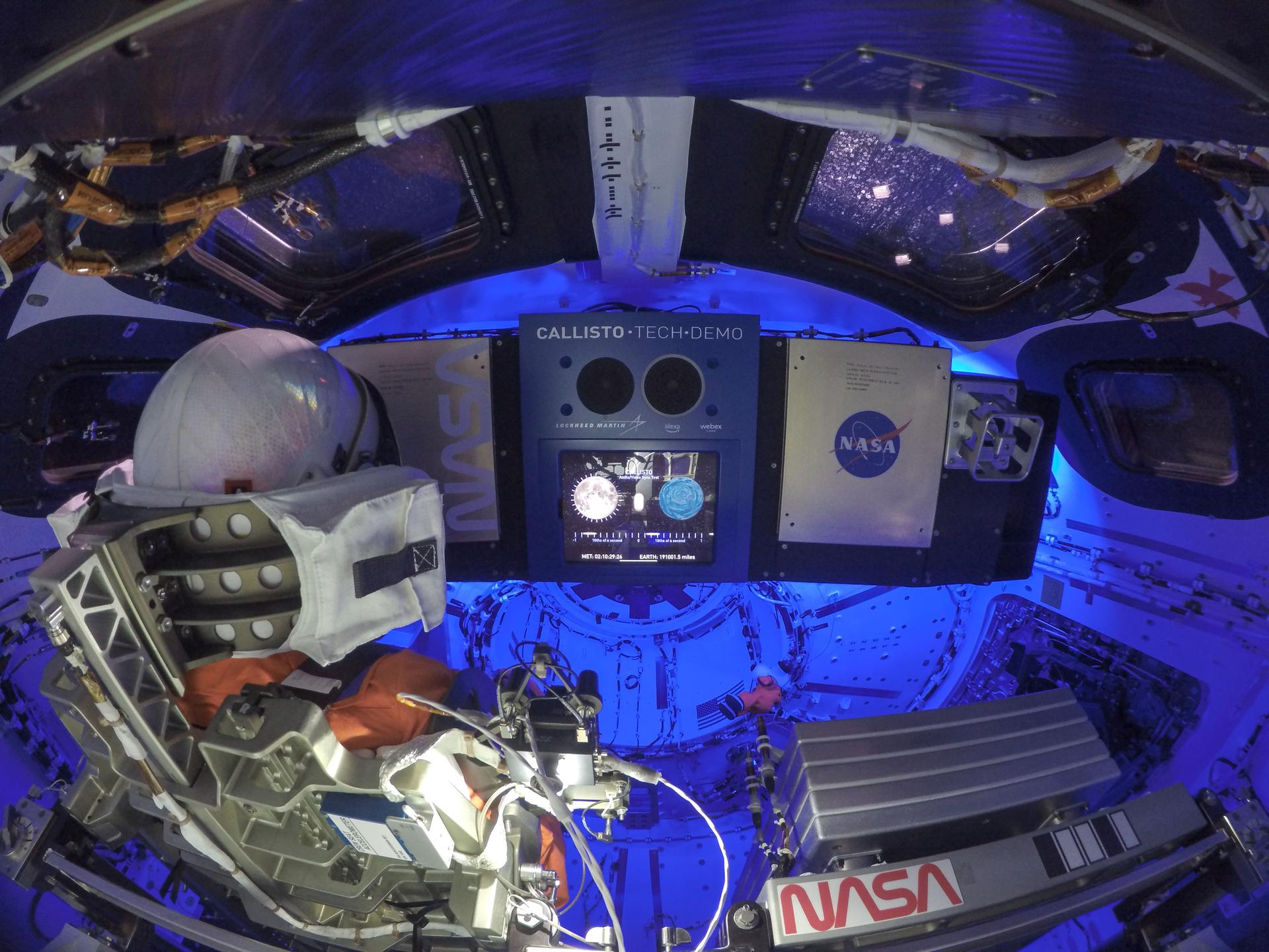 Orion's cabin interior is bathed in a blue light as the Callisto payload, center, is activated. Commander Moonikin Campos sits to the left of the photo, facing towards Callisto.