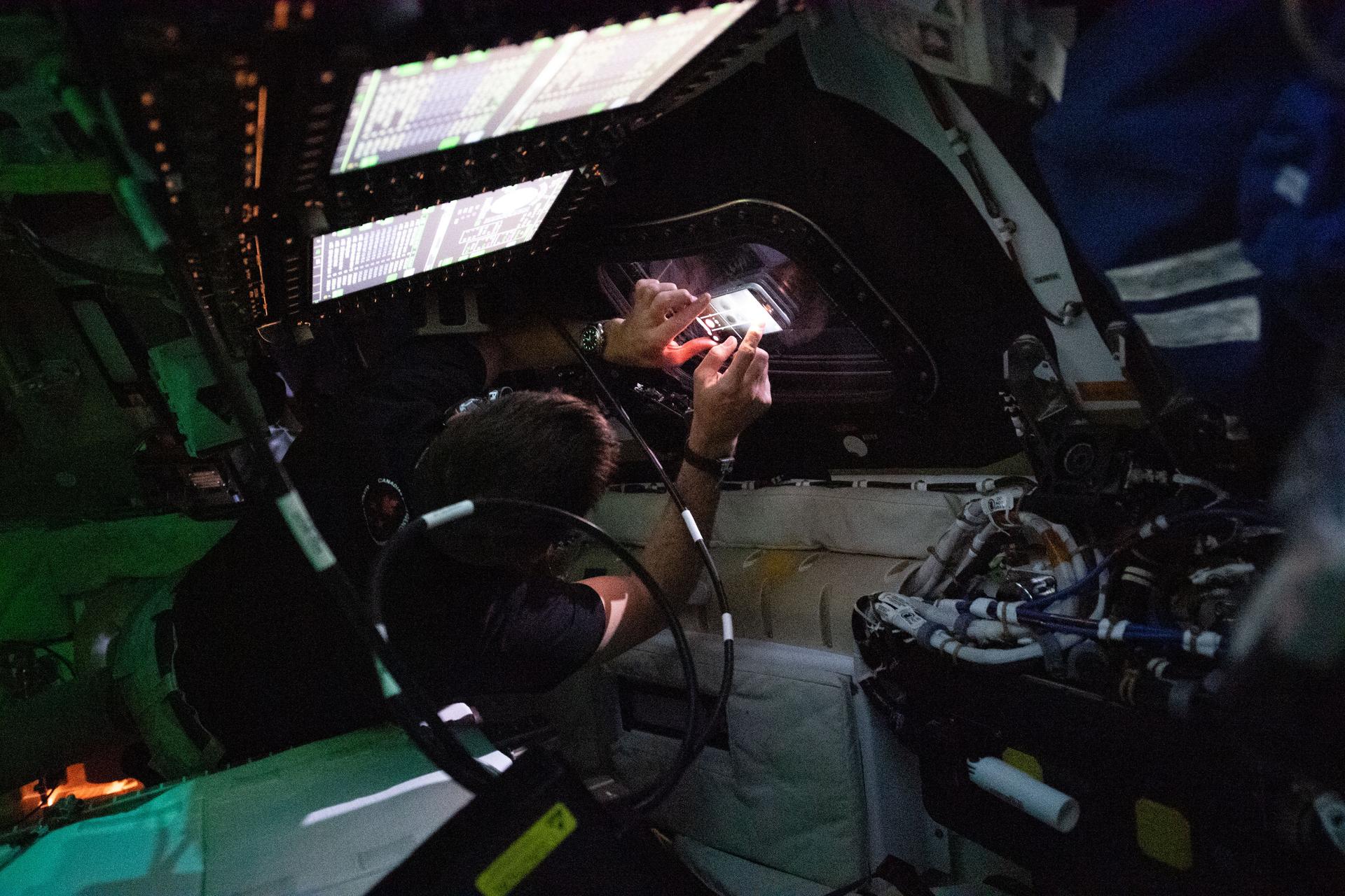 art002e025643 (April 3, 2026) - CSA (Canadian Space Agency) astronaut Jeremy Hansen – in the center of the image – about to take a photo out the window of the Orion spacecraft on day three of NASA's Artemis II mission. The controls over the commander and pilot seats are illuminated in the foreground, but the cabin is otherwise dark to avoid unnecessary glares on the windows. Credit: NASA