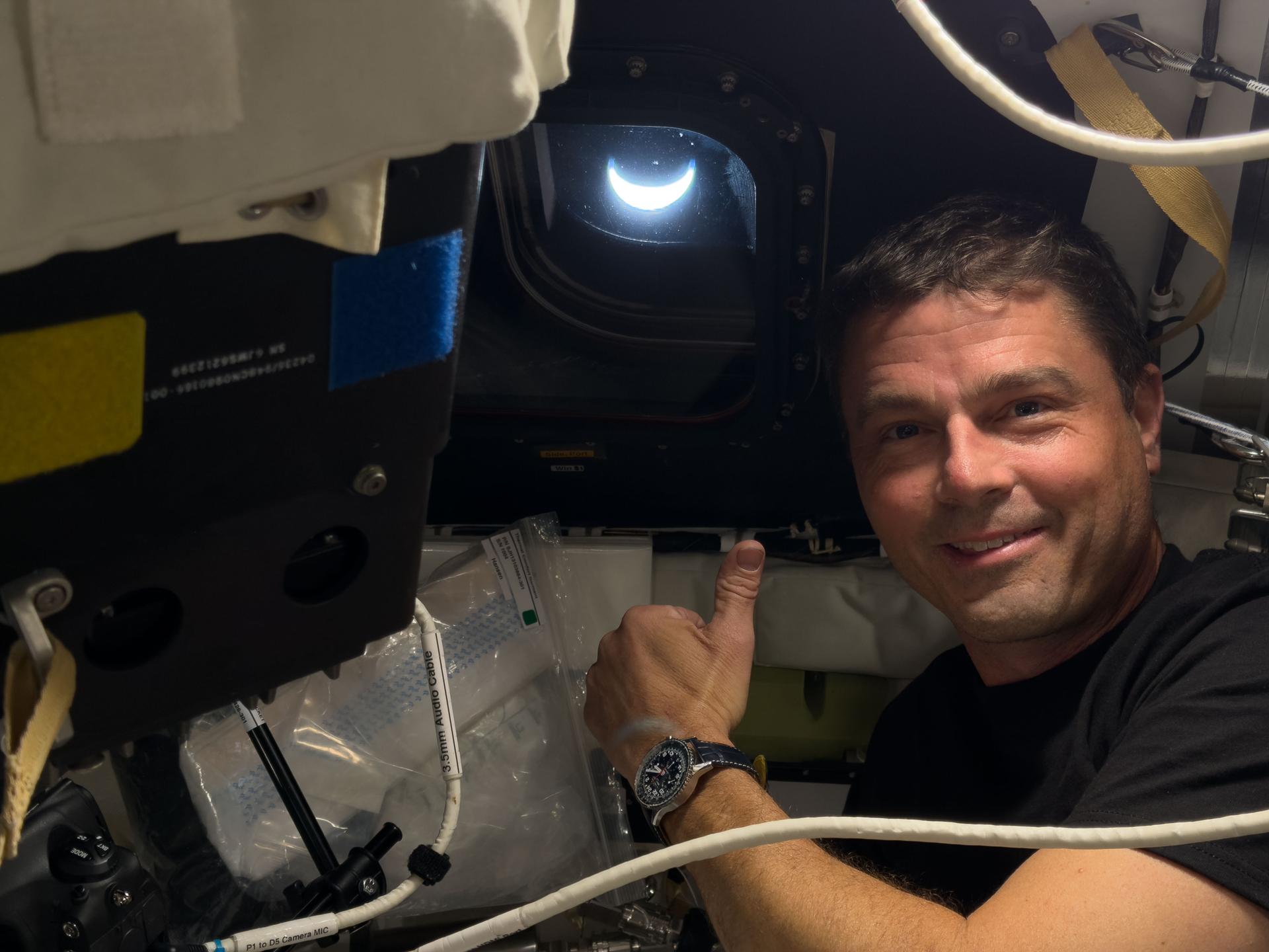 art002e041724 (April 8, 2026) - NASA astronaut and Artemis II Commander Reid Wiseman smiles aboard the Orion spacecraft with a crescent Earth visible through the window behind him on the eighth day of the Artemis II mission. Credit: NASA. Credit: NASA