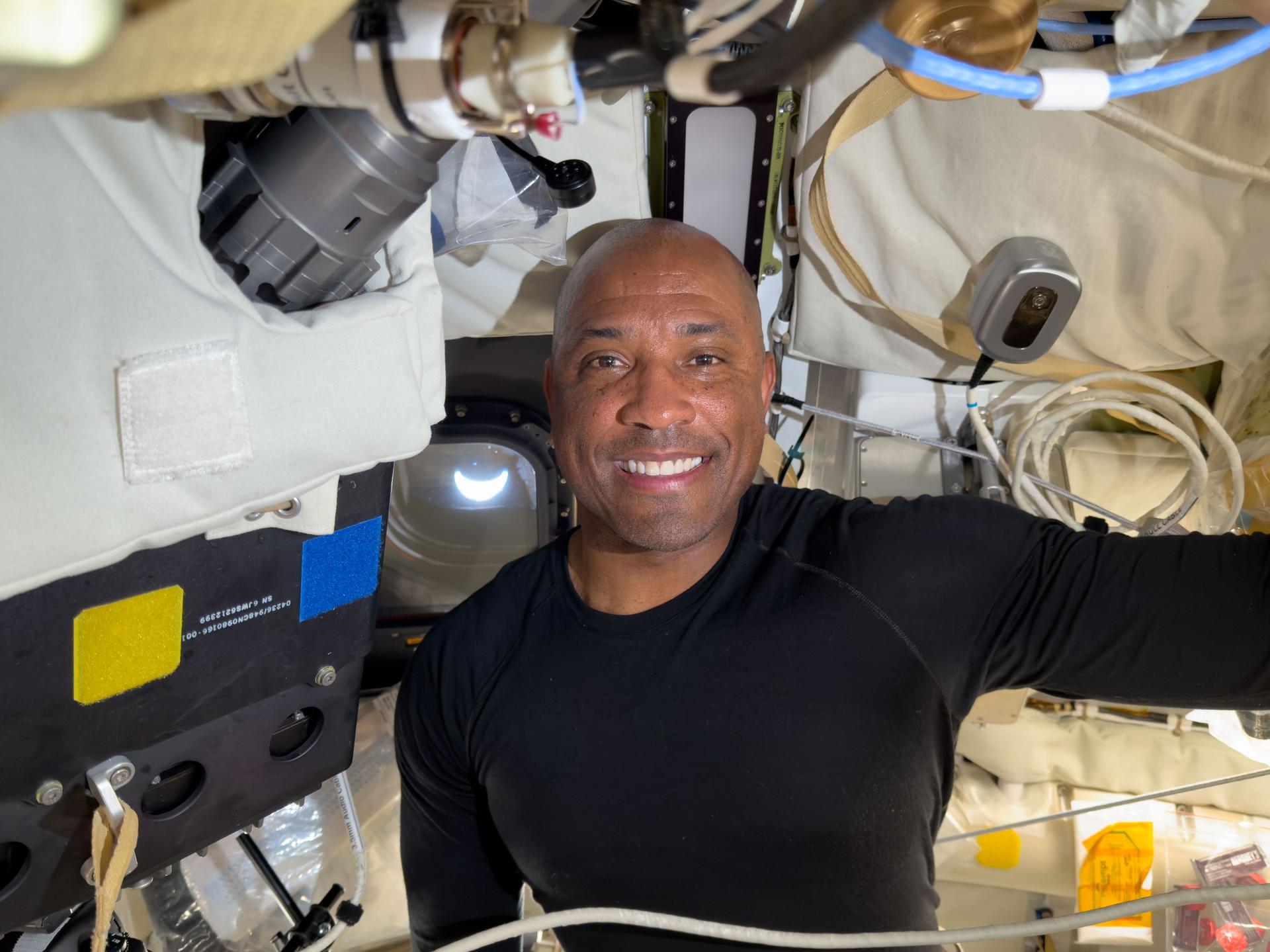 art002e041740 (April 8, 2026) - NASA astronaut and Artemis II Pilot Victor Glover smiles aboard the Orion spacecraft with a crescent Earth visible through the window behind him on the eighth day of the Artemis II mission. Credit: NASA.