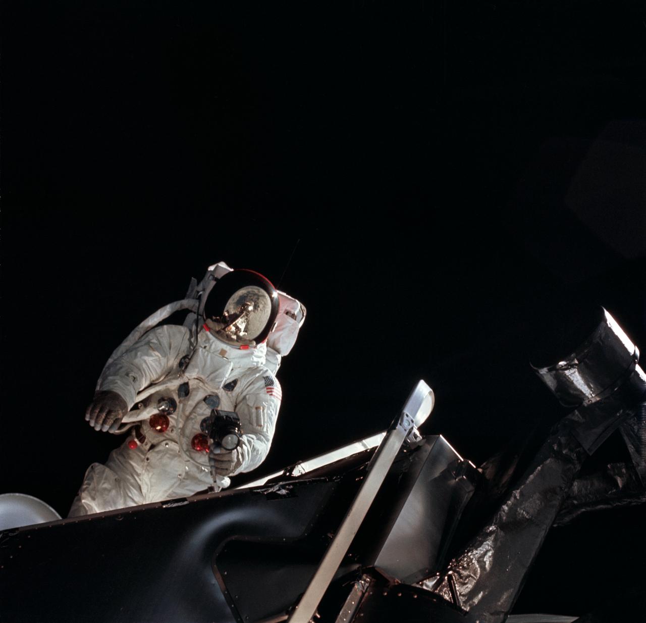 AS09-19-2983 (6 March 1969) --- Astronaut Russell L. Schweickart, lunar module pilot, operates a 70mm Hasselblad camera during his extravehicular activity (EVA) on the fourth day of the Apollo 9 Earth-orbital mission. The Command and Service Modules (CSM) and Lunar Module (LM) "Spider" are docked. This view was taken from the Command Module (CM) "Gumdrop". Schweickart, wearing an Extravehicular Mobility Unit (EMU), is standing in "golden slippers" on the LM porch. On his back, partially visible, are a Portable Life Support System (PLSS) and an Oxygen Purge System (OPS). Astronaut James A. McDivitt, Apollo 9 commander, was inside the "Spider". Astronaut David R. Scott, command module pilot, remained at the controls in the CM.