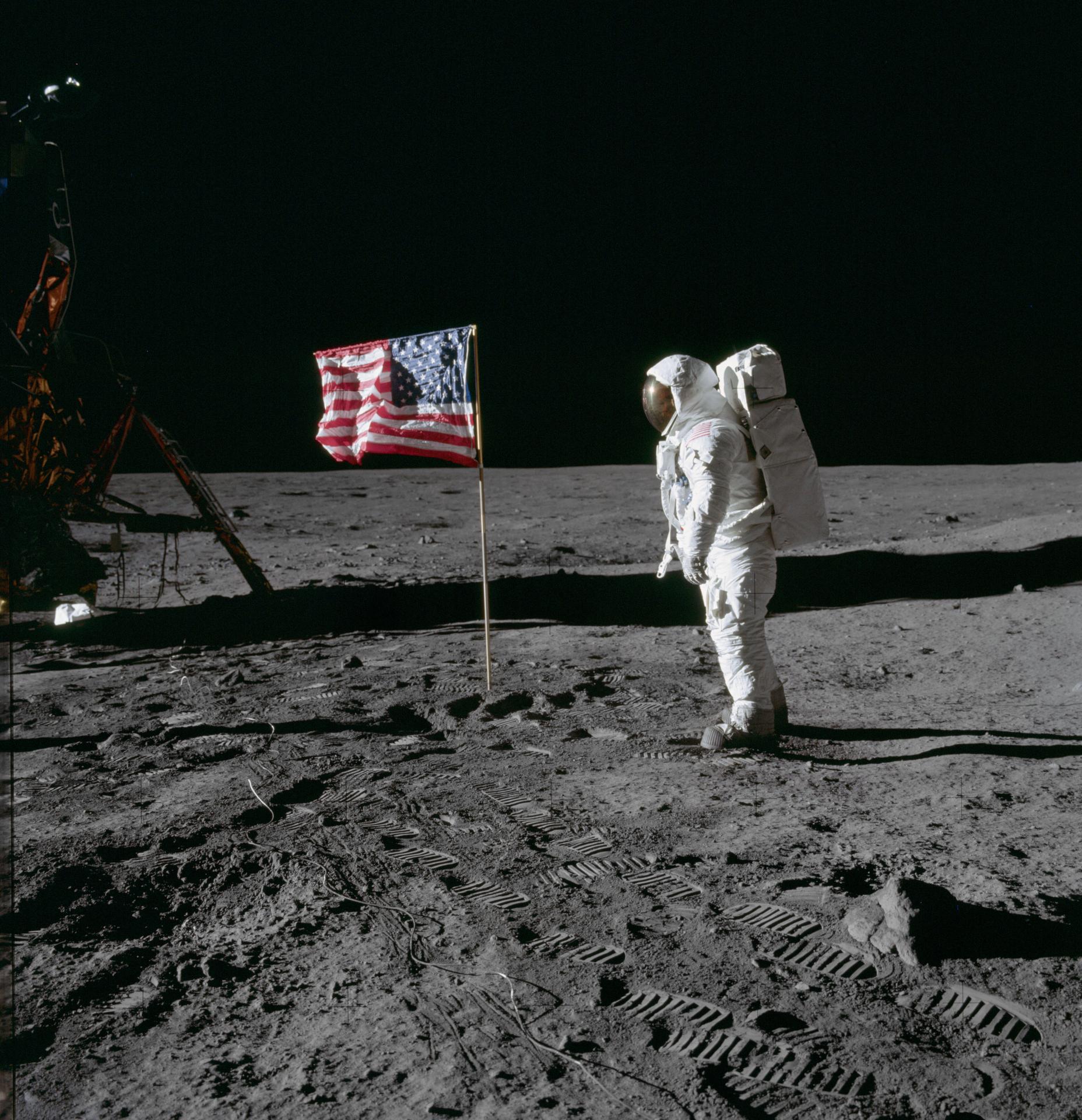 Man in a white spacesuit stands on the Moon's surface facing a U.S. flag. The side of the lunar lander is visible.