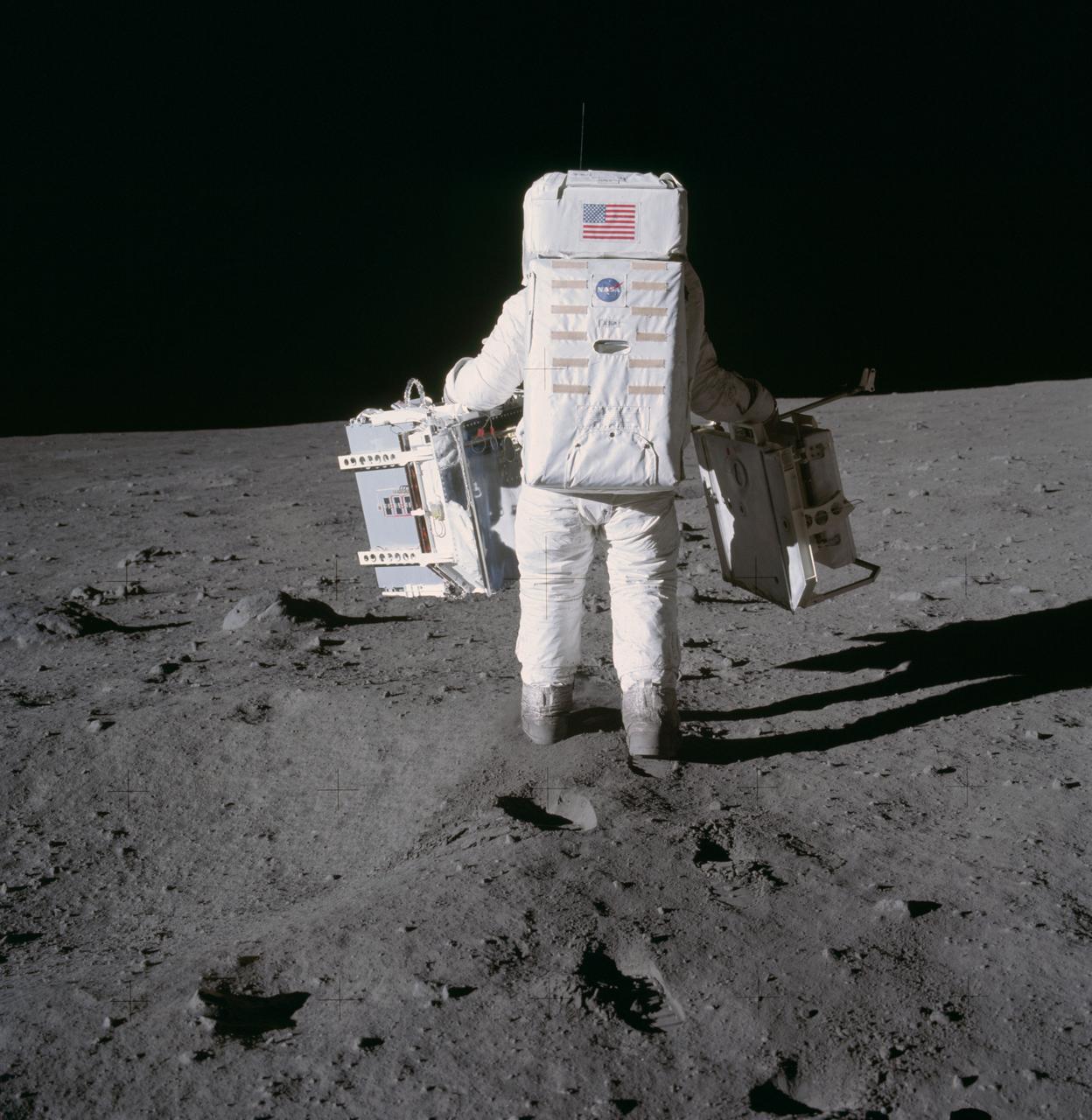 Astronaut Edwin E. Aldrin Jr., lunar module pilot, moves toward a position to deploy two components of the Early Apollo Scientific Experiments Package (EASEP) on the surface of the Moon during the Apollo 11 extravehicular activity. The Passive Seismic Experiments Package (PSEP) is in his left hand; and in his right hand is the Laser Ranging Retro-Reflector (LR3). Astronaut Neil A. Armstrong, commander, took this photograph with a 70mm lunar surface camera.