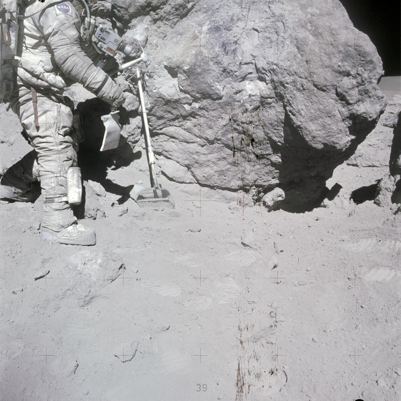 AS16-116-18653 (23 April 1972) --- Astronaut Charles M. Duke Jr., Apollo 16 lunar module pilot, stands at a big rock adjacent (south) to the huge "House Rock" (barely out of view at right edge). Note shadow at extreme right center where the two moon-exploring crew members of the mission sampled what they referred to as the "east-by-west split of House Rock" or the open space between this rock and "House Rock". At their post-mission press conference, the crewmen expressed the opinion that this rock was once a part of "House Rock" which had broken away. The two sampled the big boulder seen here also. Duke has a sample bag in his hand, and a lunar surface rake leans against the large boulder. Astronaut John W. Young, commander, exposed this view with a color magazine in his 70mm Hasselblad camera. While astronauts Young and Duke descended in the Apollo 16 Lunar Module (LM) "Orion" to explore the Descartes highlands landing site on the moon, astronaut Thomas K. Mattingly II, command module pilot, remained with the Command and Service Modules (CSM) "Casper" in lunar orbit.