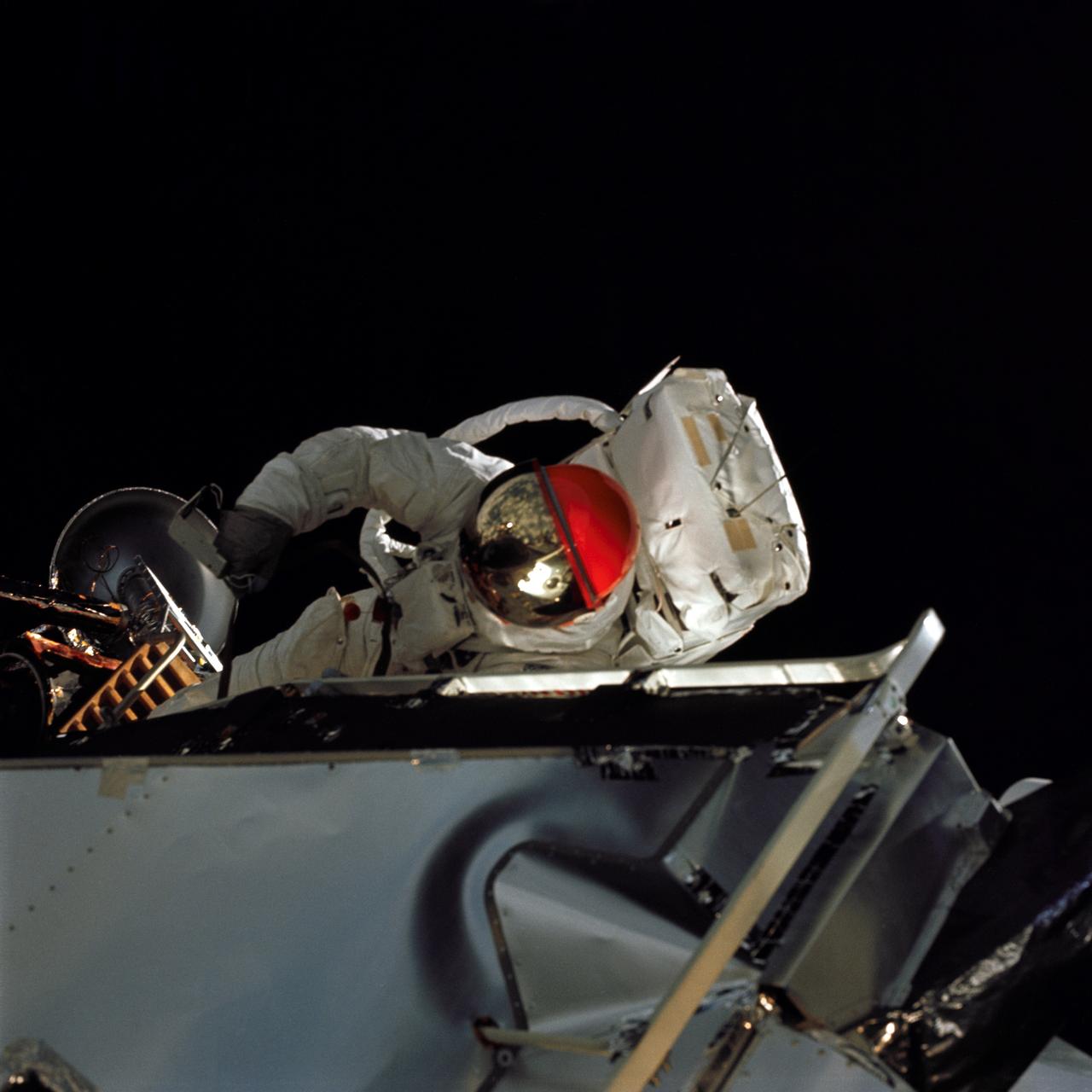 AS09-19-2994 (6 March 1969) --- Astronaut Russell L. Schweickart, lunar module pilot, is photographed from the Command Module (CM) "Gumdrop" during his extravehicular activity (EVA) on the fourth day of the Apollo 9 Earth-orbital mission. He holds, in his right hand, a thermal sample which he is retrieving from the Lunar Module (LM) exterior. The Command and Service Modules (CSM) and LM "Spider" are docked. Schweickart, wearing an Extravehicular Mobility Unit (EMU), is standing in "golden slippers" on the LM porch. Visible on his back are the Portable Life Support System (PLSS) and Oxygen Purge System (OPS). Astronaut James A. McDivitt, Apollo 9 commander, was inside the "Spider". Astronaut David R. Scott, command module pilot, remained at the controls in the CM "Gumdrop".