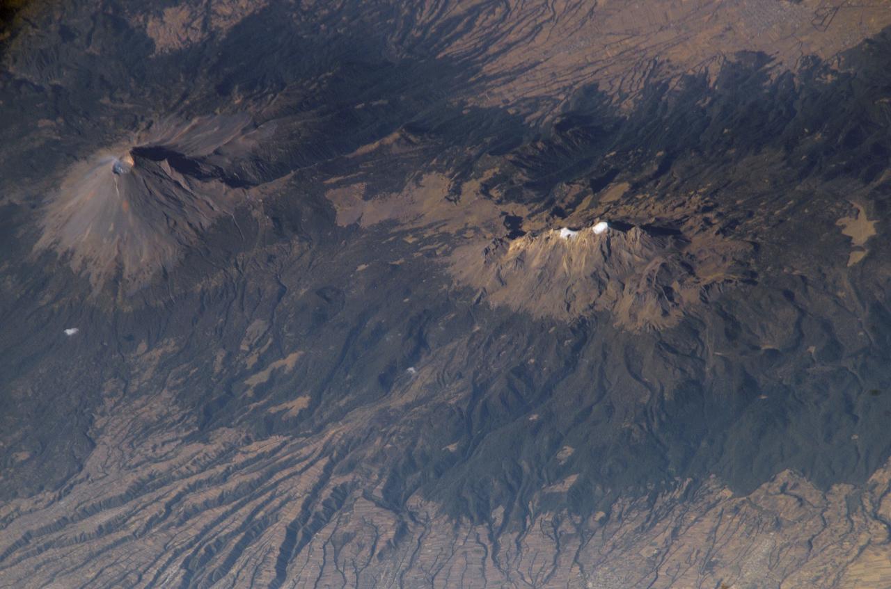ISS006-E-28546 (16 February 2003) --- Popocatepetl and Iztaccíhuatl Volcanoes in Mexico are featured in this digital image photographed by an Expedition 6 crewmember on the International Space Station (ISS). As part of the circum-Pacific “Ring of Fire”, Mexico hosts several of the world’s most continually active volcanoes including the massive Popocatepetl (Aztec for “Smoking Mountain”). This detailed oblique photograph also depicts a neighboring volcano, Iztaccíhuatl (the “Woman in White”). Popocatepetl has produced small, intermittent eruptions since 1994. In addition to the constant potential of eruptions producing ash deposits, pyroclastic flows, and lava, the summit of Popocatepetl also hosts glaciers. These can melt during eruptions to form mudflows that blanket areas to the south. In contrast to Popocatepetl’s well-defined symmetrical cone, Iztaccíhuatl is formed from several overlapping smaller cones that trend north-northwest to south-southeast. Glaciers and year-round snow are also present on Iztaccíhuatl (white regions along the peaks). According to NASA scientists, deep valleys have been eroded into the massive apron of ash and pumice deposits, glacial outwash, and alluvium to the east of the volcano. Despite its close proximity, similar age, and similar geologic character to Popocatepetl, Iztaccíhuatl has not erupted in historic times. This has encouraged the establishment of numerous agricultural fields (visible as faint rectilinear patterns in the lower half of the image) on the eastern flank of the mountain.