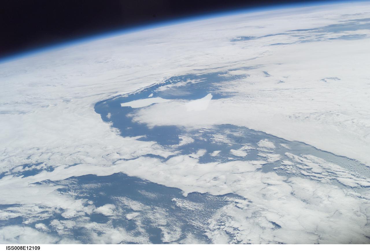 ISS008-E-12109 (6 January 2004) --- Five year old icebergs near South Georgia Island are featured in this image photographed by an Expedition 8 crewmember onboard the International Space Station (ISS). This oblique image shows two pieces of a massive iceberg that broke off from the Antarctica Ronne Ice Shelf in October 1998. The pieces of iceberg A-38 have floated relatively close to South Georgia Island. After five years and 3 months, they are approximately 1500 nautical miles from their origin. The cloud pattern is indicative of the impact of the mountainous islands on the local wind field. At the time this image was taken, the icebergs were sheltered in the lee side of the island.