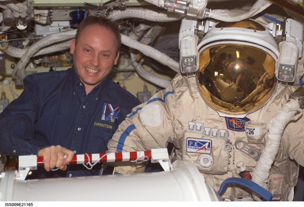 ISS009-E-21165 (1 September 2004) --- Astronaut Edward M. (Mike) Fincke, Expedition 9 NASA ISS science officer and flight engineer, poses with his Russian Orlan spacesuit in the Pirs Docking Compartment of the International Space Station (ISS). The container for the pump panel is in the foreground.