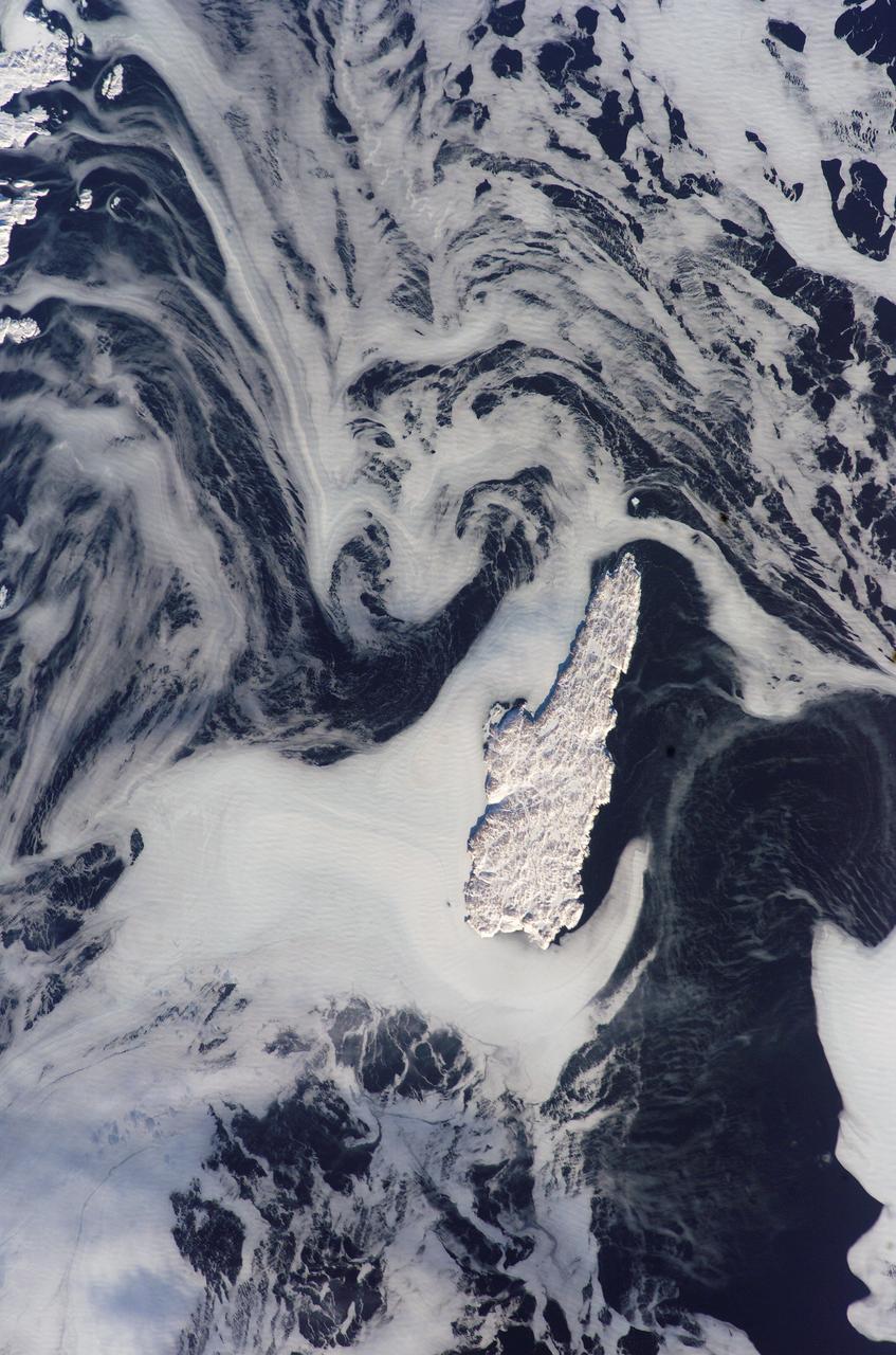 ISS012-E-15918 (24 Jan. 2006) --- Belle Isle and a portion of Newfoundland, Canada are featured in this image photographed by an Expedition 12 crew member on the International Space Station. Belle Isle (center) is surrounded by sea ice in this recent winter view. Belle Isle lies in the strait between the Island of Newfoundland and Labrador (the mainland portion of Canada&#0146;s province of Newfoundland). Small islands along the coast of Labrador appear in the top left corner. In this key location Belle Isle lies on the shortest shipping lanes between the Great Lakes and Europe, and also on the main north-south shipping route to Hudson Bay and the Northwest Territories. Snow and ice in this recent winter view obliterate the dozens of glacier-scoured lakes that dot the surface of the island. The single community of Belle Isle Landing on the southeast tip is equally hard to see. Ice patterns also show that the island lies at the meeting point of two sea currents. The Labrador Current flows from the northwest (top left), and a smaller current, driven by dominant westerly winds, flows from the southwest (lower left) out of the narrow Belle Isle Strait (out of frame lower left). Flow lines in sea ice indicate the sense of movement of the ice.  Ice floes embedded in the Labrador Current appear in the upper part of the image as a relatively open pattern. Sea ice with a denser pattern enters from the lower left corner, banking against the west side of Belle Isle. Tendrils flow around capes at either end of the island, with an ice-free &#0147;shadow&#0148; on the opposite, downstream side. Eddies (center) in the ice patterns show where the currents interact, north and west of the island.