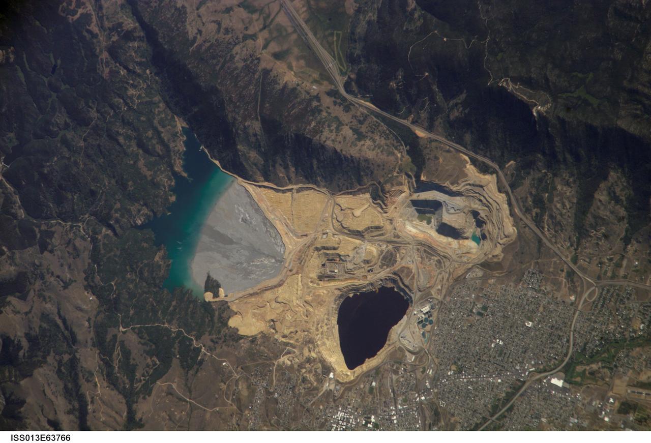ISS013-E-63766 (2 Aug. 2006) --- Berkeley Pit and Butte, Montana are featured in this image photographed by an Expedition 13 crewmember on the International Space Station. The city of Butte, Montana has long been a center of mining activity. Underground mining of copper began in Butte in the 1870s, and by 1901 underground workings had extended to the groundwater table. Thus began the creation of an intricate complex of underground drains and pumps to lower the groundwater level and continue the extraction of copper. Water extracted from the mines was so rich in dissolved copper sulfate that it was also "mined" (by chemical precipitation) for the copper it contained. In 1955, the Anaconda Copper Mining Company began open-pit mining for copper in what is now know as the Berkeley Pit (dark oblong area in center). The mine took advantage of the existing subterranean drainage and pump network to lower groundwater until 1982, when the new owner ARCO suspended operations at the mine. The groundwater level swiftly rose, and today water in the Pit is more than 900 feet deep. Many features of the mine workings are visible in this image such as the many terraced levels and access roadways of the open mine pits (gray and tan sculptured surfaces). A large gray tailings pile of waste rock and an adjacent tailings pond are visible to the north of the Berkeley Pit. Color changes in the tailings pond are due primarily to changing water depth. The Berkeley Pit is listed as a federal Superfund site due to its highly acidic water, which contains high concentrations of metals such as copper and zinc. The Berkeley Pit receives groundwater flowing through the surrounding bedrock and acts as a "terminal pit" or sink for these heavy metal-laden waters. Ongoing efforts include regulation of water flow into the pit to reduce filling of the Pit and potential release of contaminated water into local aquifers or surface streams.