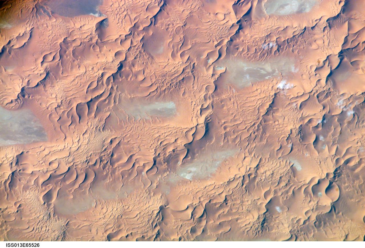 ISS013-E-65526 (8 Aug. 2006) --- Issaouane Dune Sea, Eastern Algeria is featured in this image photographed by an Expedition 13 crewmember on the International Space Station. This view from one of the smaller dune seas in the central Sahara shows the complex but regular patterns produced by winds in deserts where abundant sand is available. Geologists now know that dune seas (also called ergs) comprise at least three orders of dune size. In this image the largest and oldest appear here as chains oriented about 60 degrees apart, that is, one oriented almost north-south, the other southwest-northeast.  The "streets" between the dune chains (also called mega-dunes) are swept clean of sand in places, revealing the original surface, with light colored muds and salt derived from very occasional rains. The chains have probably taken hundreds of thousands of years to accumulate, starting when the Sahara began to become significantly dry roughly 2.5 million years ago.  Rivers became smaller, failed to reach the sea and deposited their sand load in the desert.  Wind did the rest, blowing the sand into aerodynamic dune forms. According to scientists, chain trends coincide with two of the four major trends identified in the Great Eastern Sand Sea immediately to the north. Each trend likely implies a different formative wind direction--attesting to the climate shifts that have occurred since sand began to accumulate in the central Sahara. Smaller dunes are superimposed on the mega-dunes. Sinuous crest lines are the mesoscale (intermediate in size) forms, forming octopus-like crests, especially evident as the arms of star dunes. Whereas the mega-dunes are apparently stationary, studies based on aerial photographs in other parts of the world show that these dune crests move in the course of decades. The smallest dunes appear in patches on the eastern sides of the mega-dunes as a tracery of closely spaced crests. Small dunes move fast and reform quickly as stronger winds shift with the seasons. Sand grains are blown continuously from upwind dunes, across the dune-free flats. Small dunes form when the grains slow down and accumulate at the next large dune. The small dunes ride up and over the backs of the mega- and meso-dunes. Interestingly the crest orientation of the small dunes is different from that of the mesoscale dunes throughout the image. This is a common effect of wind direction shifting locally depending on dune height: the increased friction caused by larger dunes causes formative winds to blow to the left of the (weaker) winds that form the small dunes. The friction effect of larger dunes is to the right in the southern hemisphere, well illustrated on the coast of the Namib Desert.